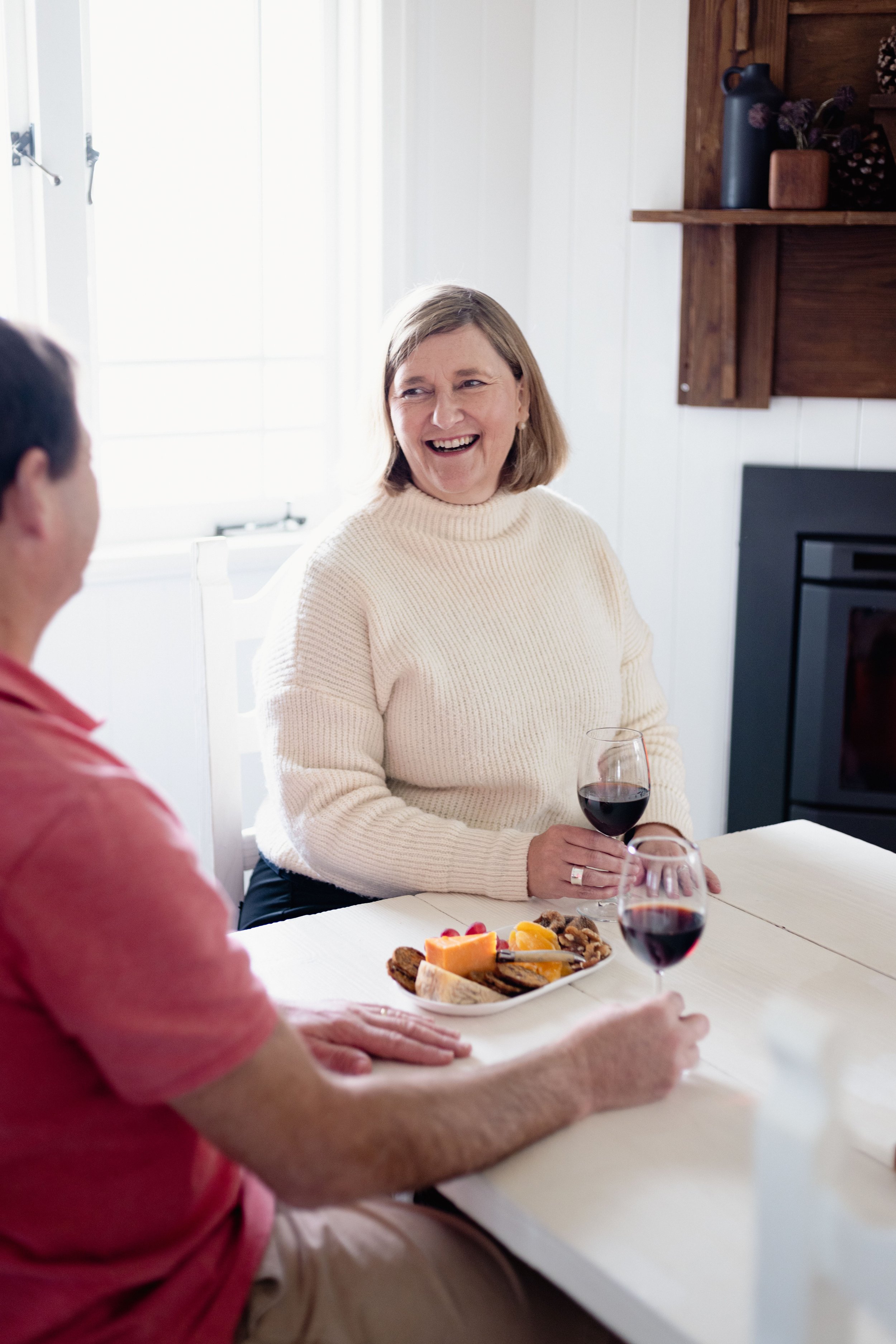 Two people at a table with wine and snacks, sharing a laugh in a cozy room."}