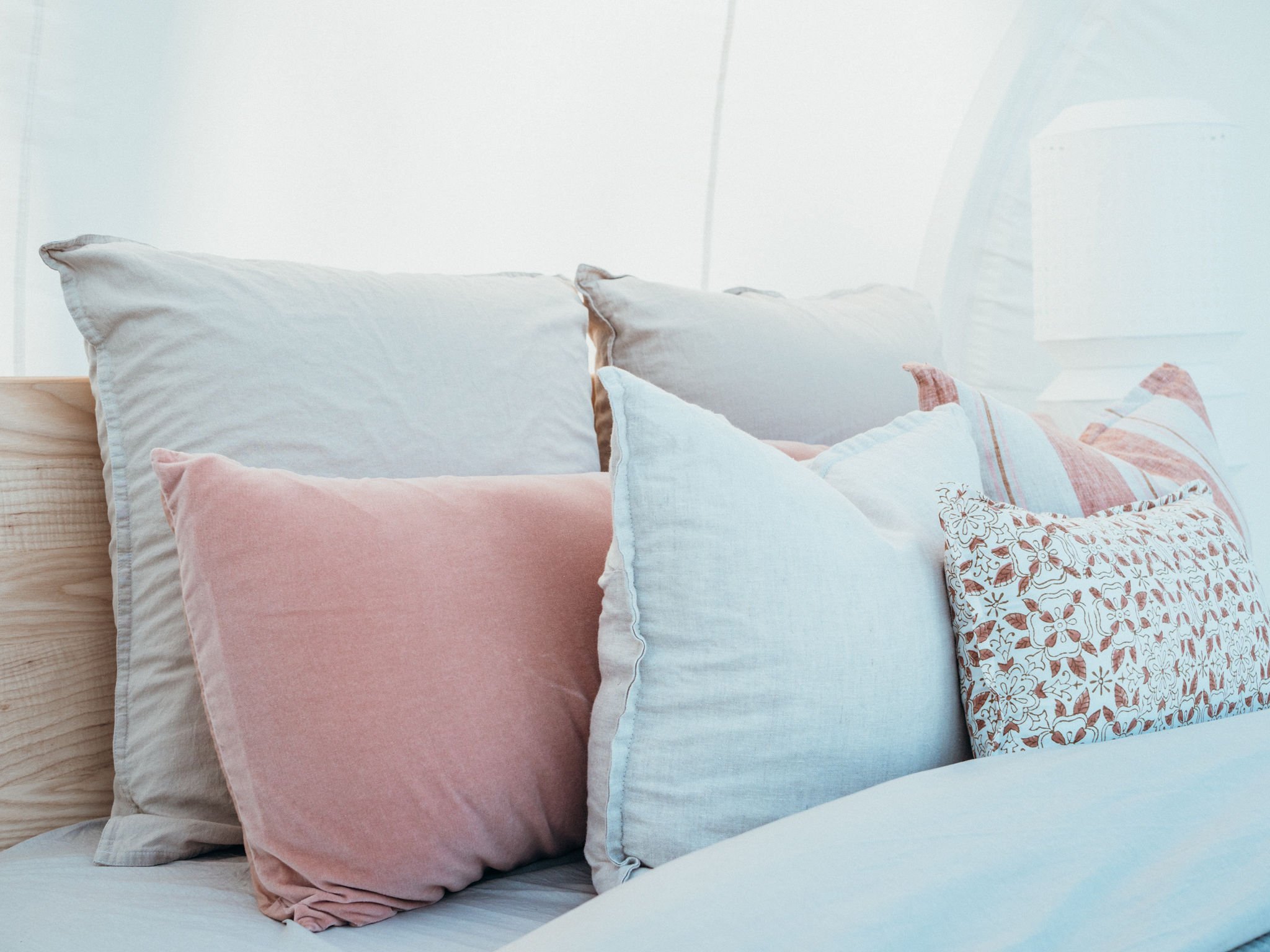 A collection of pastel-colored throw pillows arranged on a bed, featuring various textures and patterns including floral and striped designs.