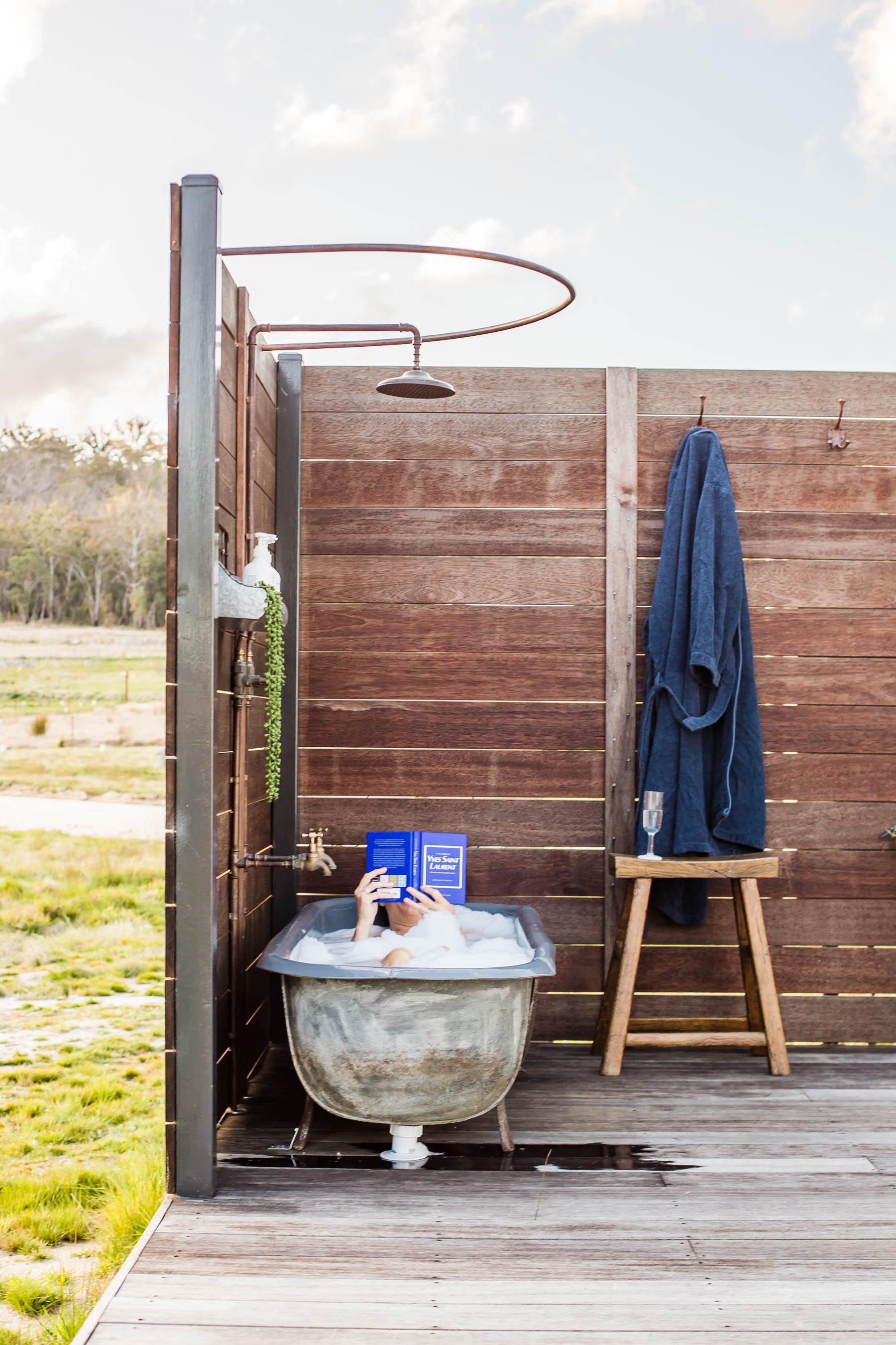 Outdoor wooden bath area with clawfoot tub, person reading a book, towel on a hook, and a stool with a glass.