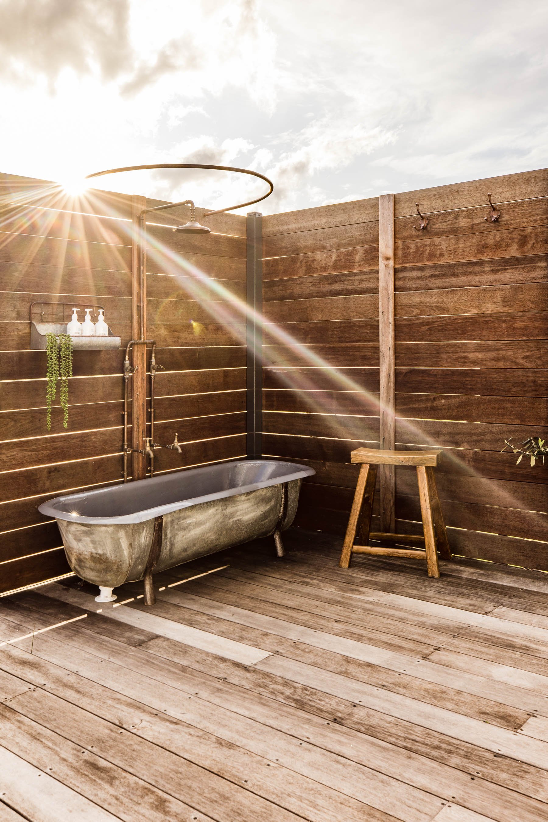 Outdoor rustic bathroom with freestanding metal bathtub, wooden walls, wooden stool, sunlight streaming through, and hanging shelf with bottles and plants.