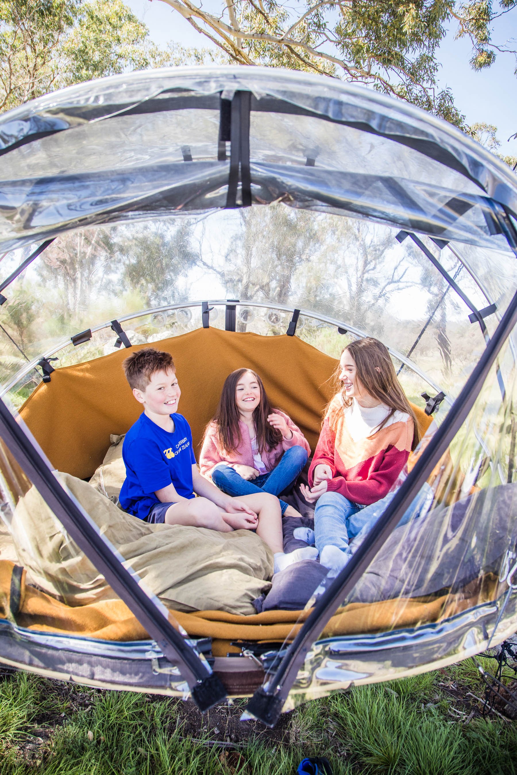 Three kids sitting and chatting inside a transparent Tiny Bubble tent, surrounded by trees and grass in a wooded area.