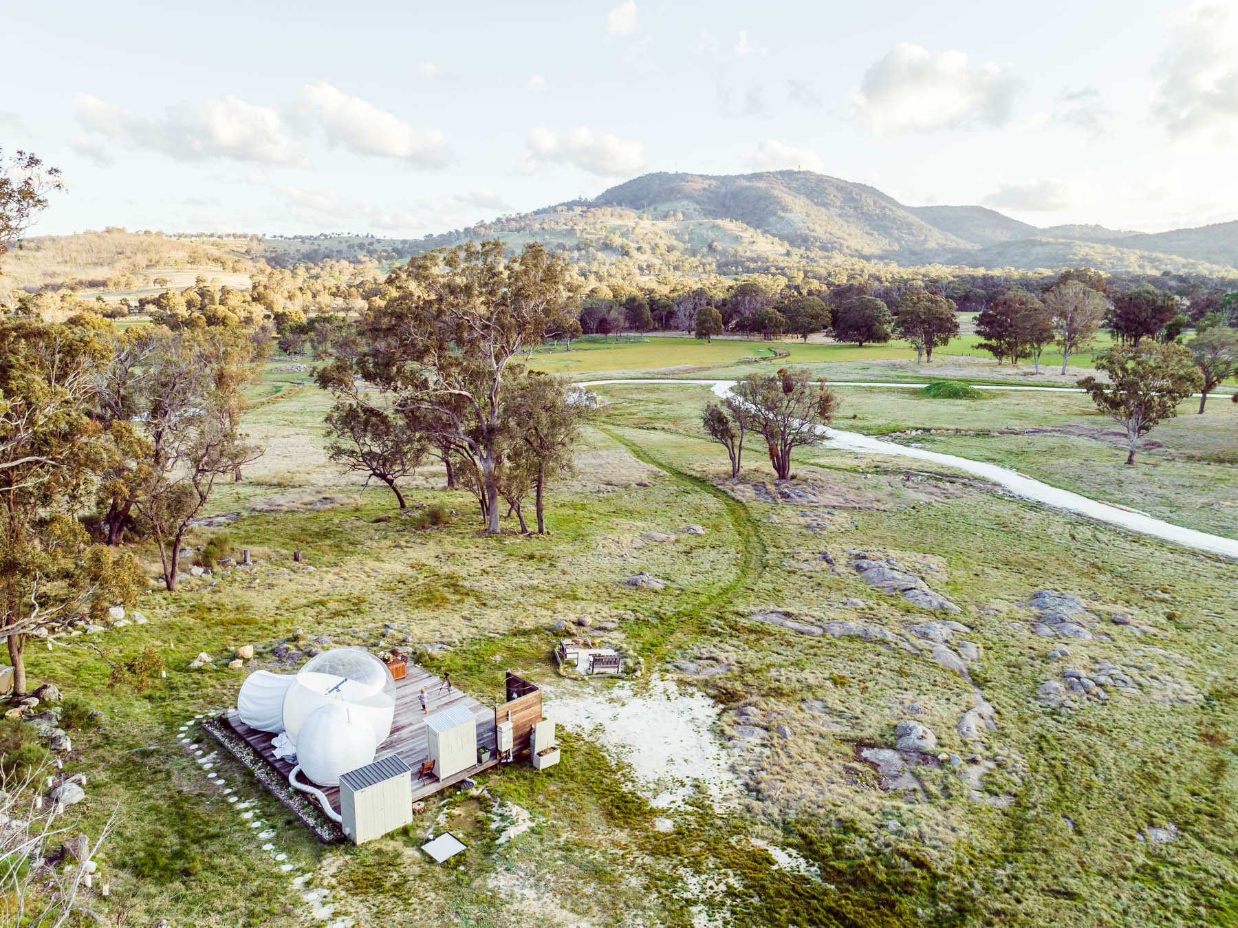 Scenic landscape with small rustic cabin and inflatable bubble tent in a grassy field, surrounded by trees and hills in the background.