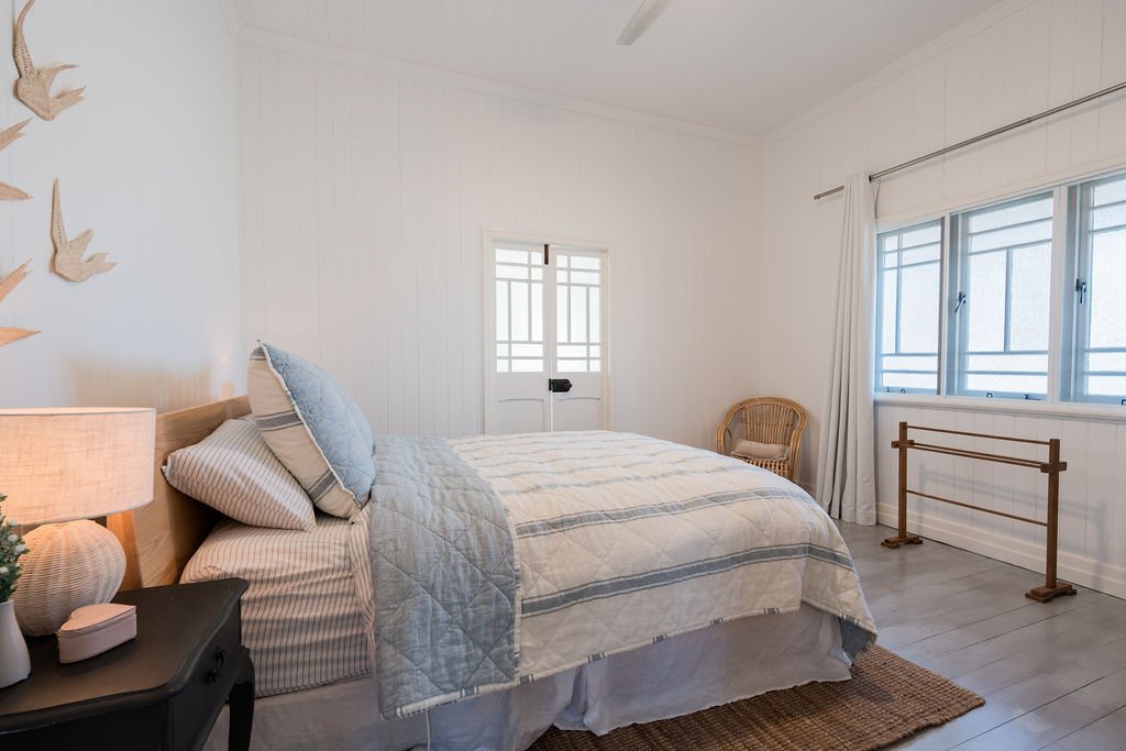 Minimalist bedroom with a light color palette, featuring a bed with striped pillows and quilt, a wicker chair, a nightstand with a lamp, wall art of birds, and a window with white curtains.