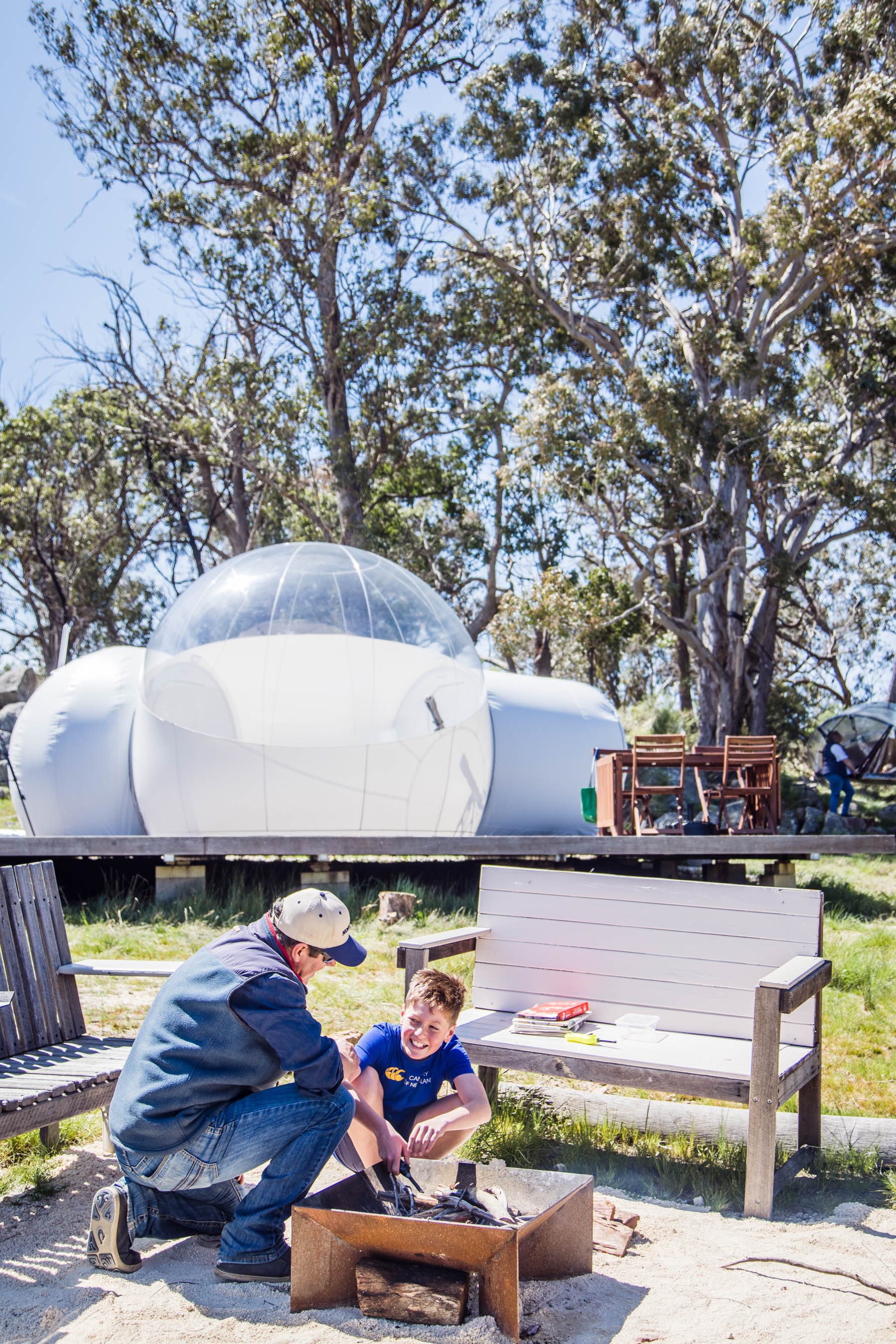 Man and boy at campsite with a fire pit, Bubbletent, and outdoor seating area with trees in the background.