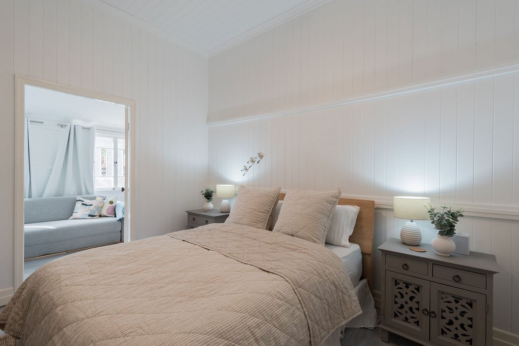 Cozy bedroom with beige bedding and pillows, grey bedside tables with lamps, and potted plants, leading into a living area with a light grey sofa and white curtains.
