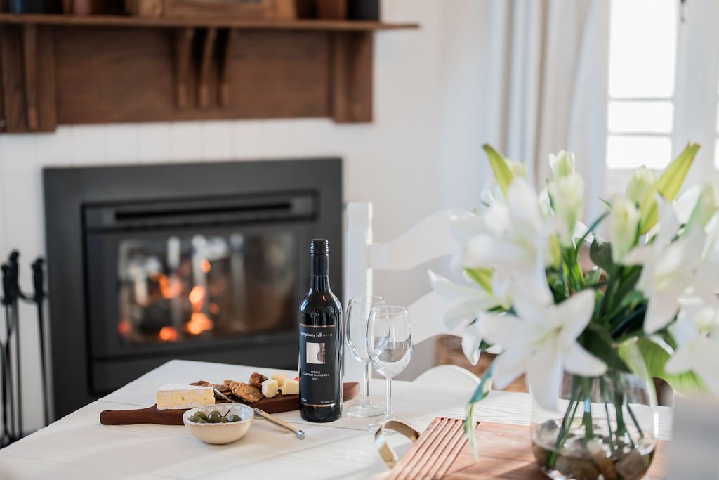Cozy dining setup with a bottle of red wine, wine glass, plate of bread and cheese, bowl of olives, white flowers, and a lit fireplace in the background.