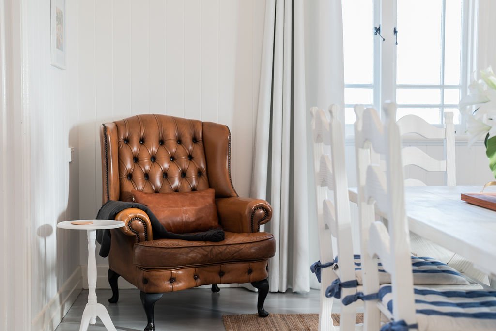 Cozy living space with a brown leather tufted armchair, white side table, and white wooden dining chairs with blue striped cushions. Light gray curtains and a bright window in background, creating a warm, inviting atmosphere.