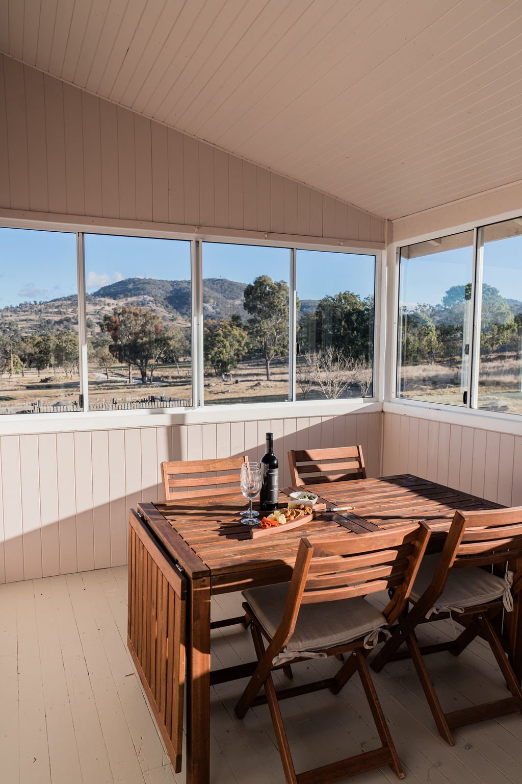 Indoor patio with a wooden table set for two, wine bottle, glass, and charcuterie board; large windows with scenic mountain and tree view.