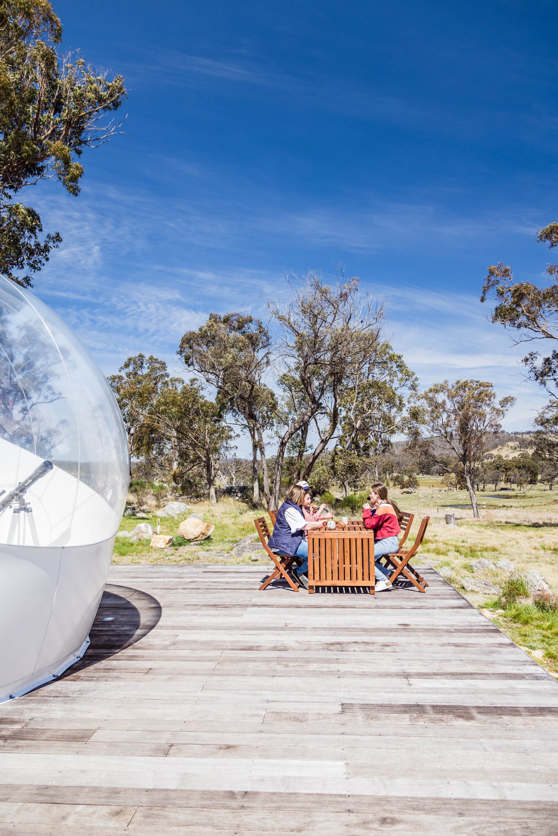 Two people sitting at a wooden table on a deck next to a Bubbletent, surrounded by trees and open field under a clear blue sky.