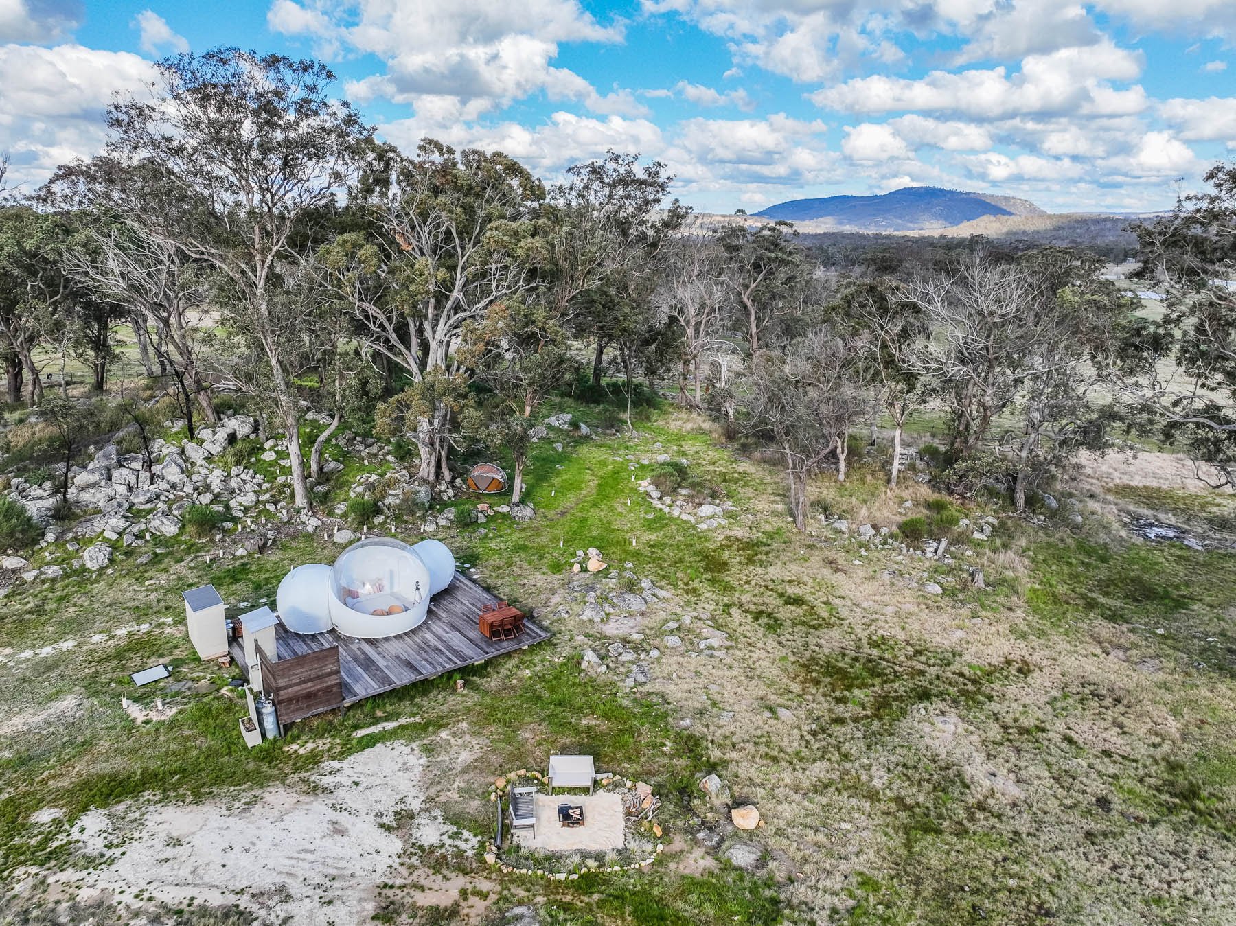 Aerial view of a glamping setup in a natural setting with trees and rocks. Features include a Bubbletent and a wooden deck. Scenic landscape with grassy fields, scattered stones, and a distant mountain under a blue sky with clouds.