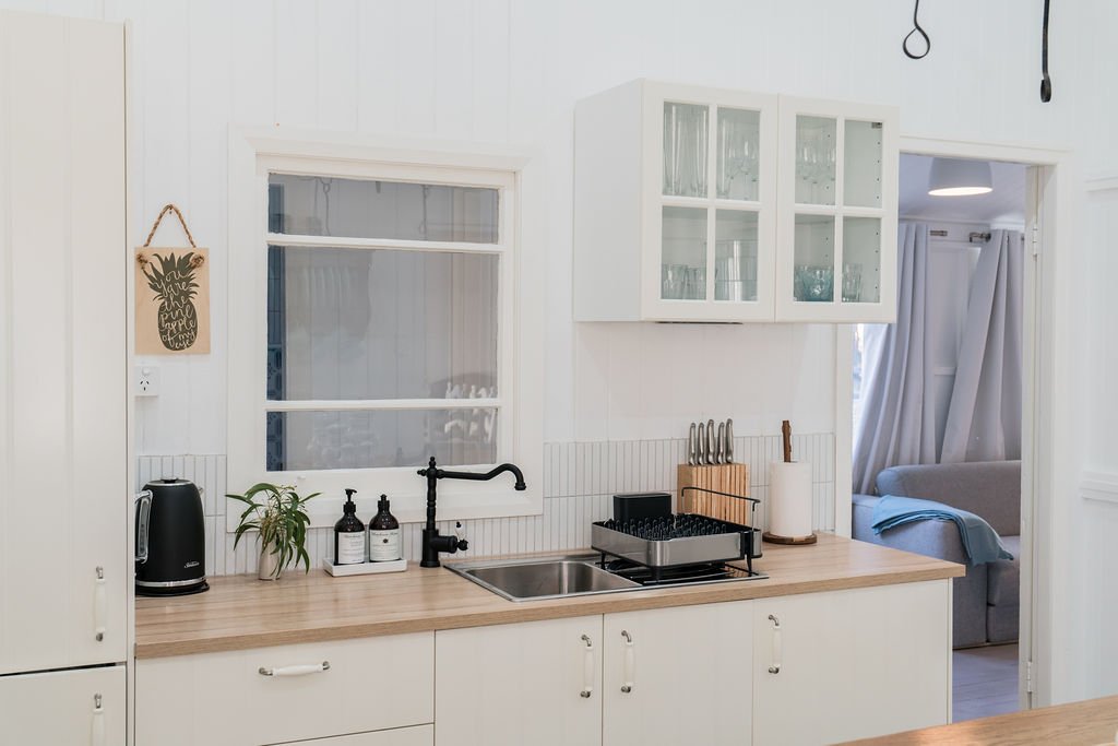 Modern kitchen with wooden countertop, stainless steel sink, black , and utensils. White cabinets with glass doors above, a black kettle, plant, soap bottles, knife block, and dish rack. Sign with pineapple artwork on wall. Living room visible throug