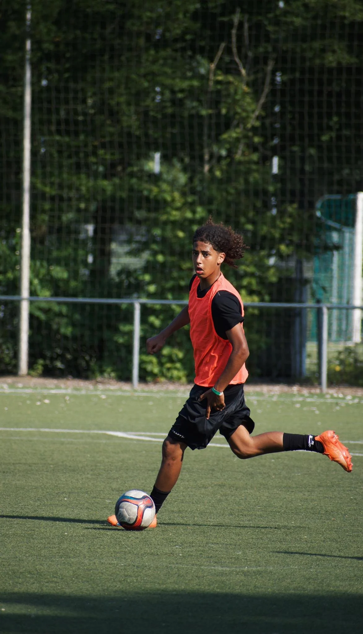 A young male soccer player in black shorts, black shoes, and a red jersey kicking a soccer ball on a green field, with a fence and trees in the background.
