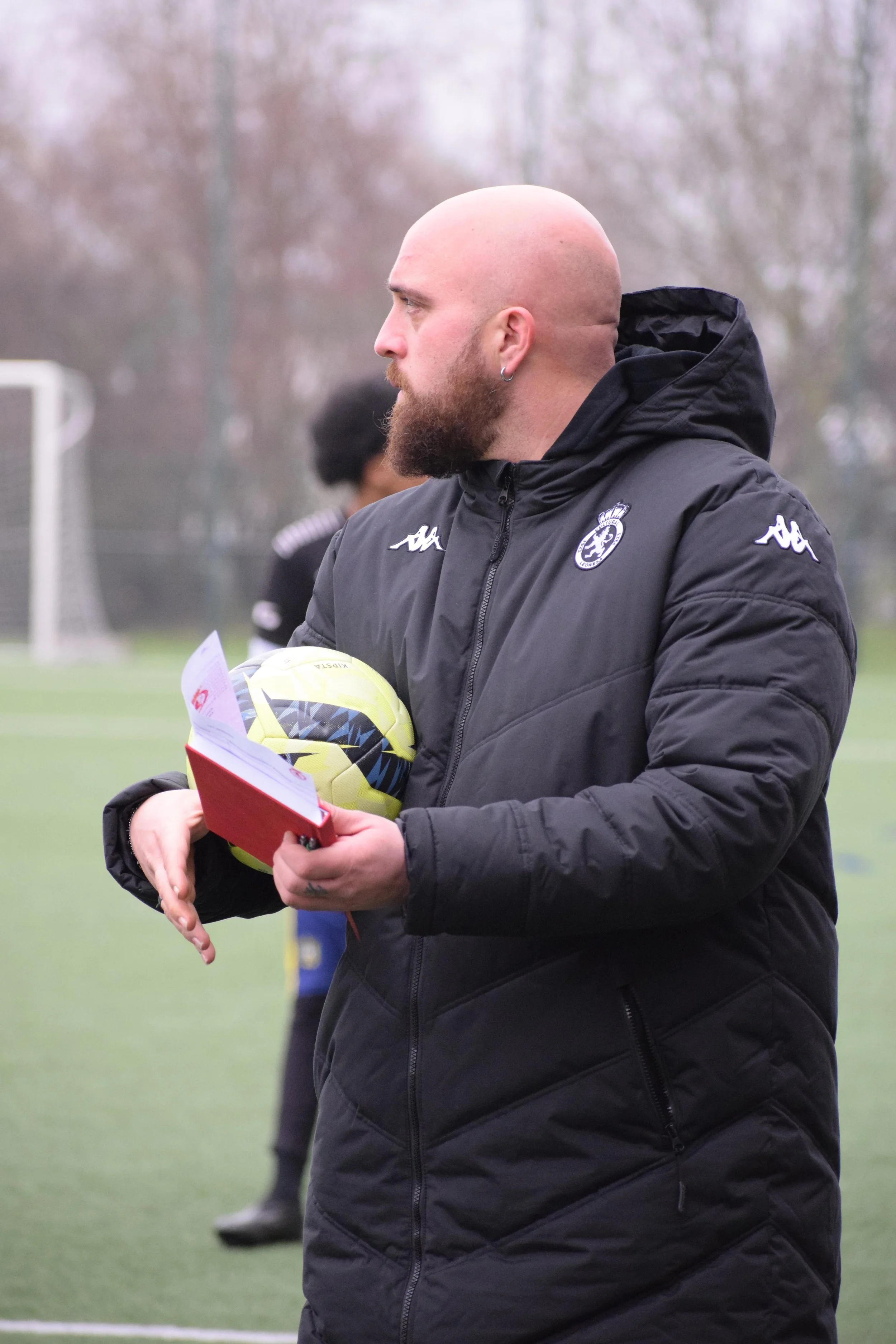 A bald man with a beard, wearing a black jacket with a football club logo, holding a yellow soccer ball and a red notebook, standing on a soccer field.