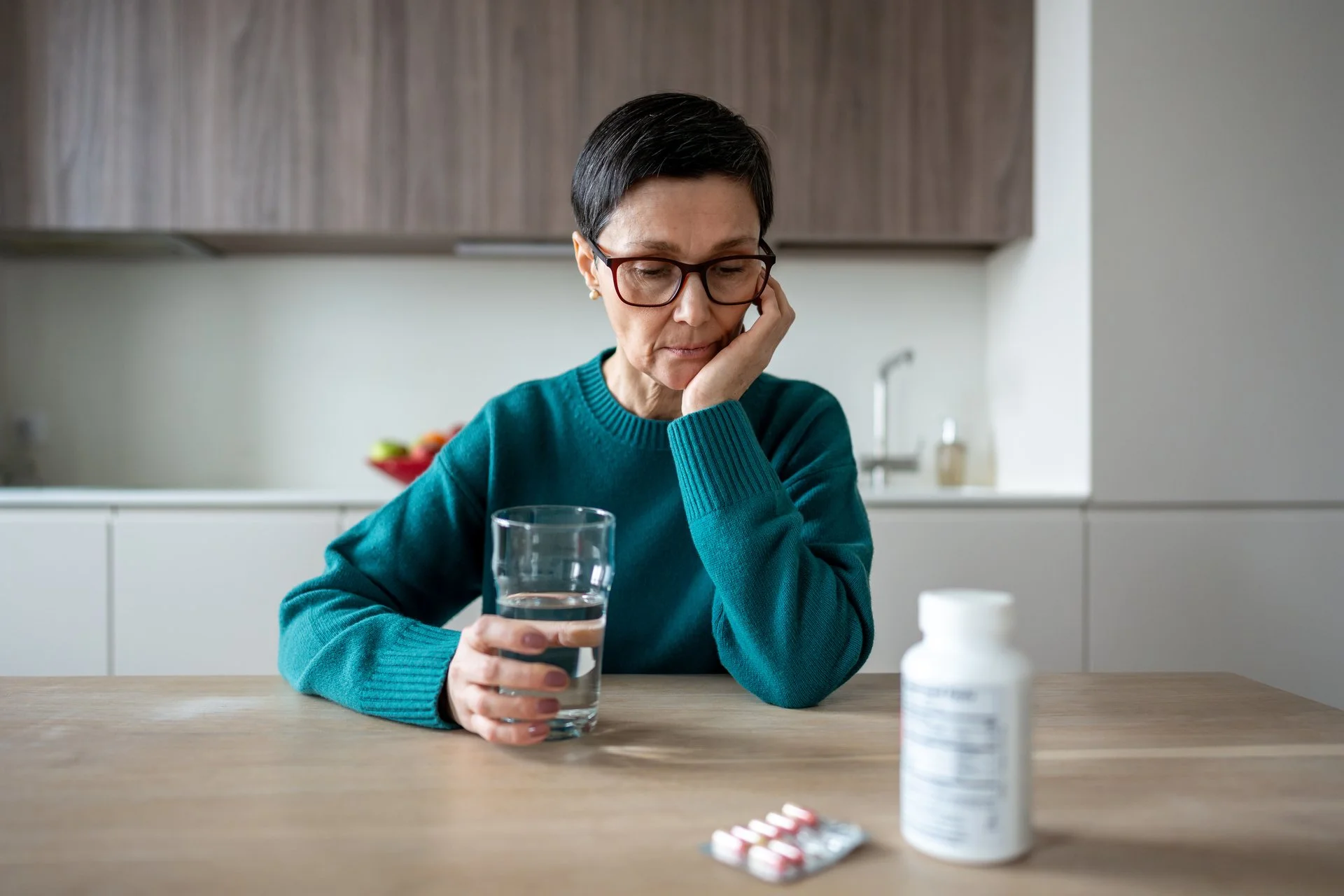 Woman sitting at a table with a pensive look on her face and a bottle of pills sitting in front of her on the table.