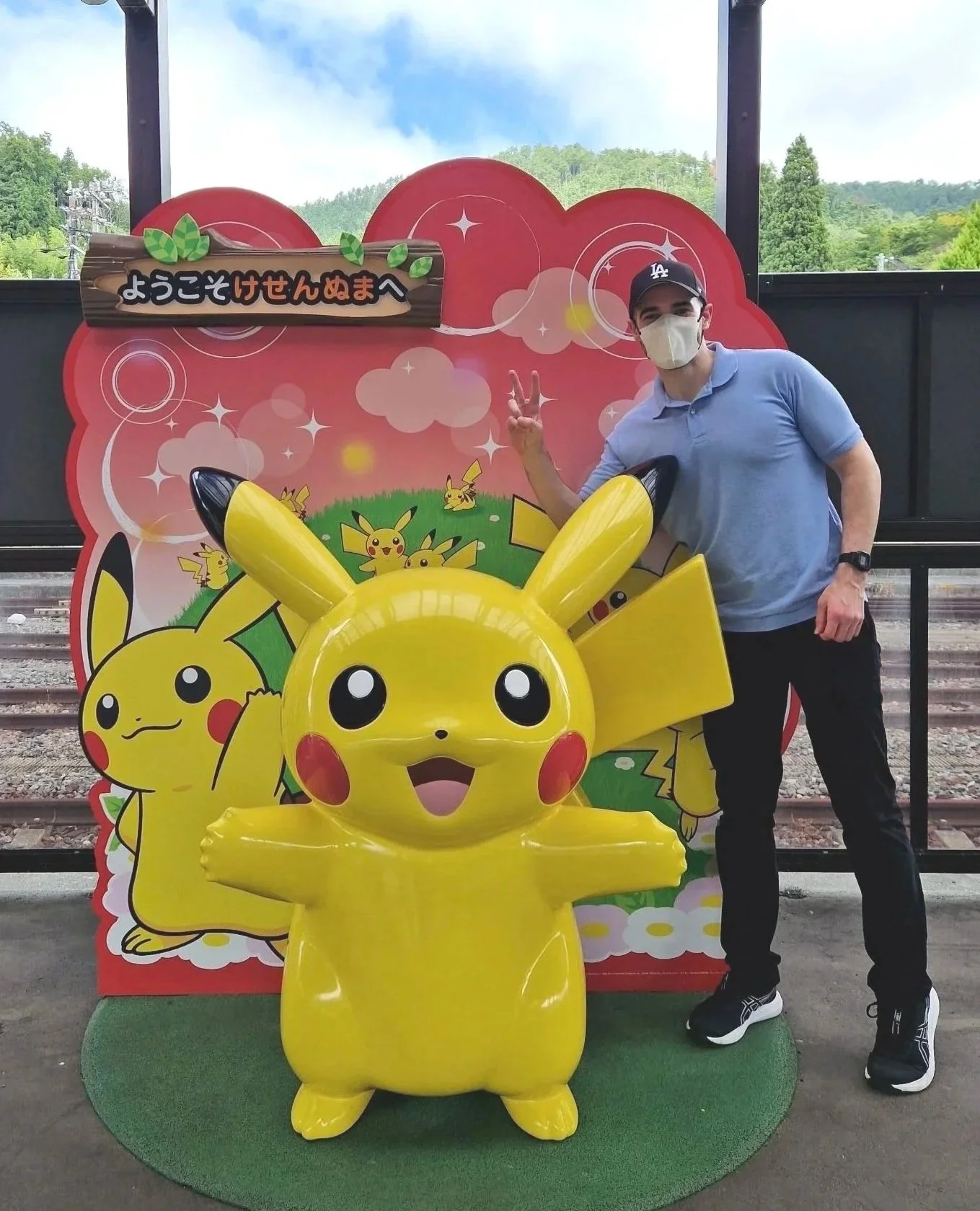 A man wearing a face mask, baseball cap, and casual clothing standing next to a large Pikachu figure at a Pokémon-themed location with a red backdrop and green grass floor.