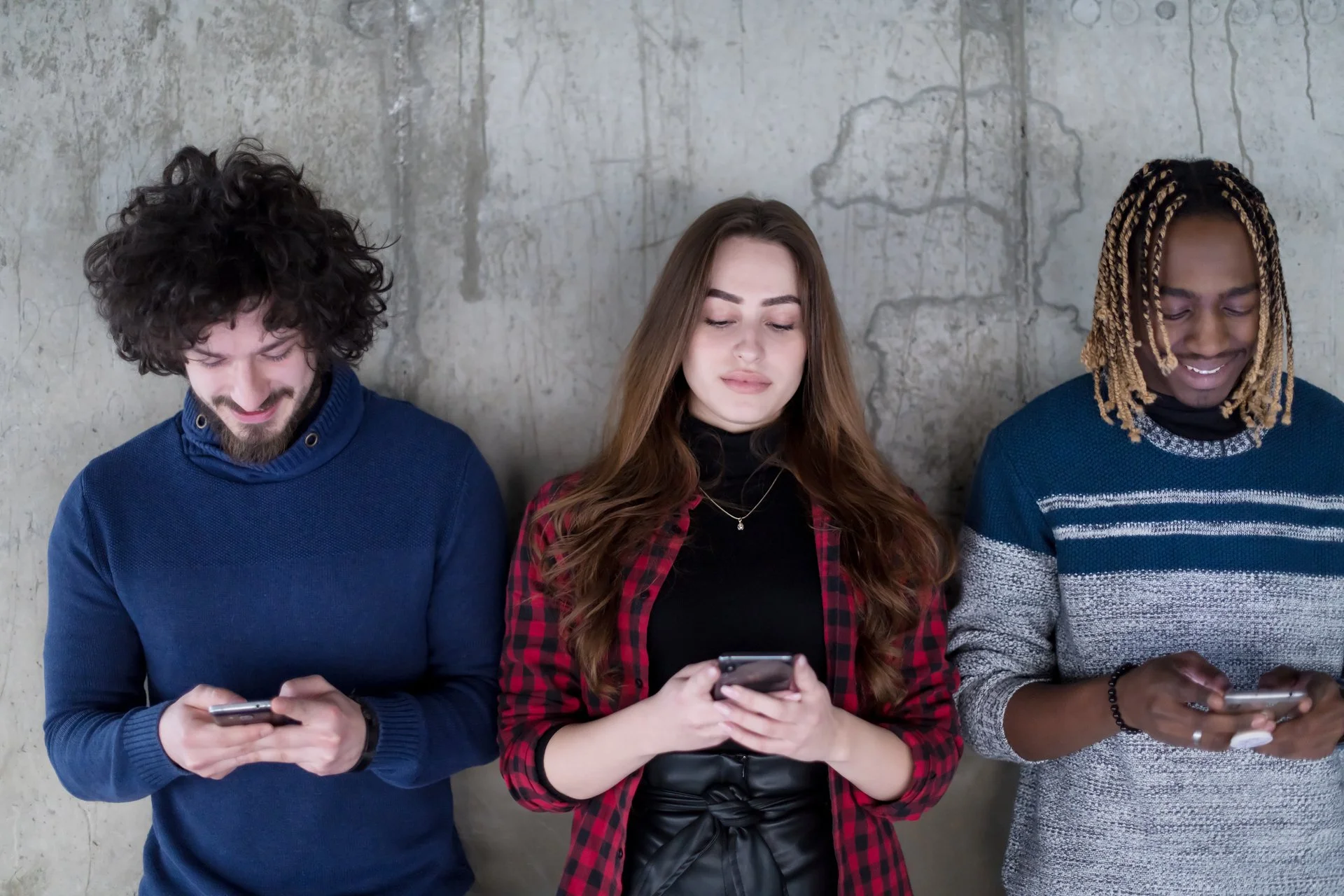 A group of three people standing side-by-side but not interacting with one another because they are on their phones.