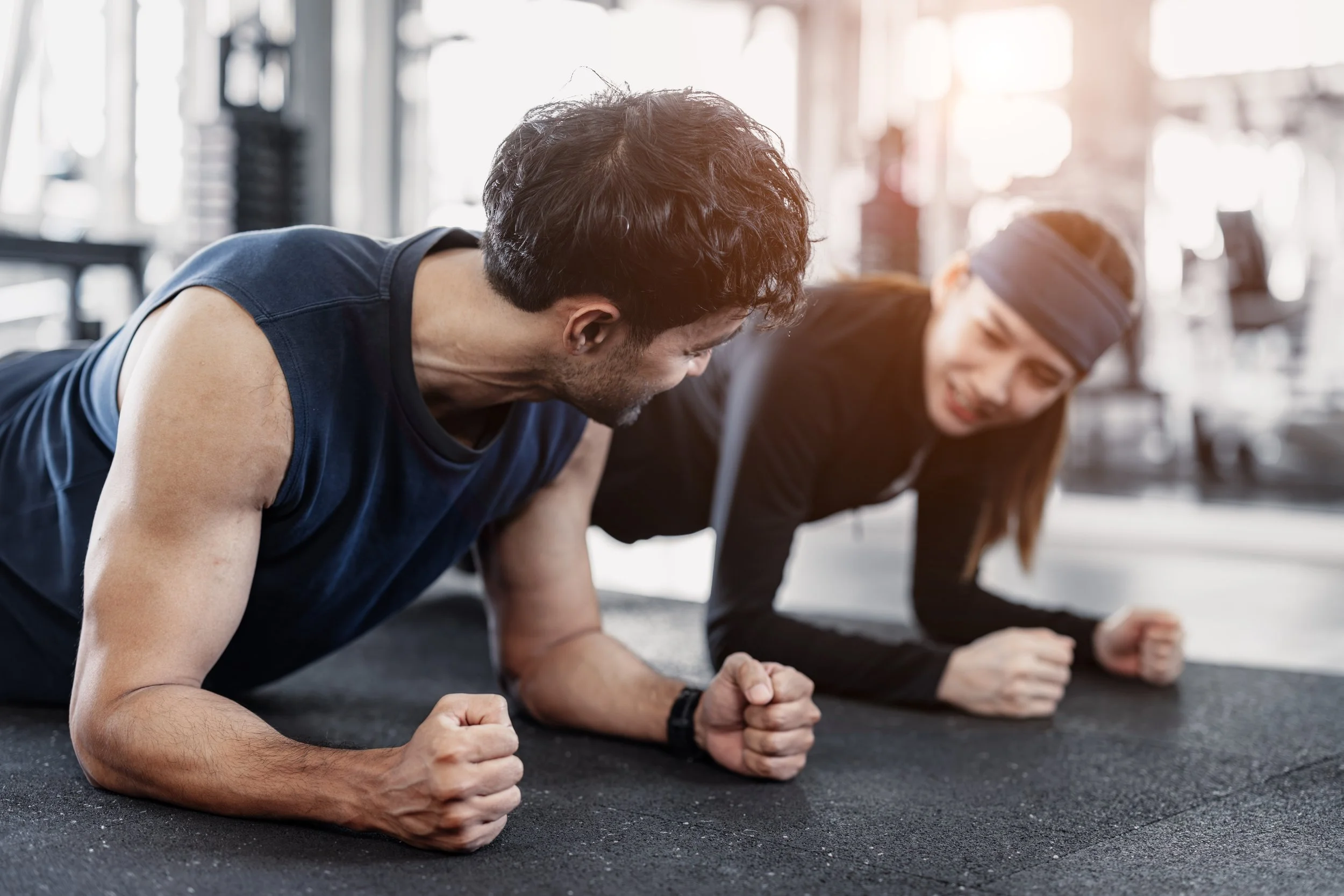 Man and a woman planking side by side in a gym.