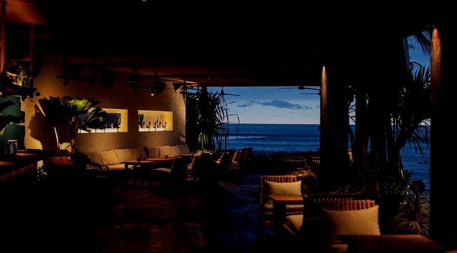 Dimly lit tropical lounge area with outdoor seating and a view of the ocean during sunset.