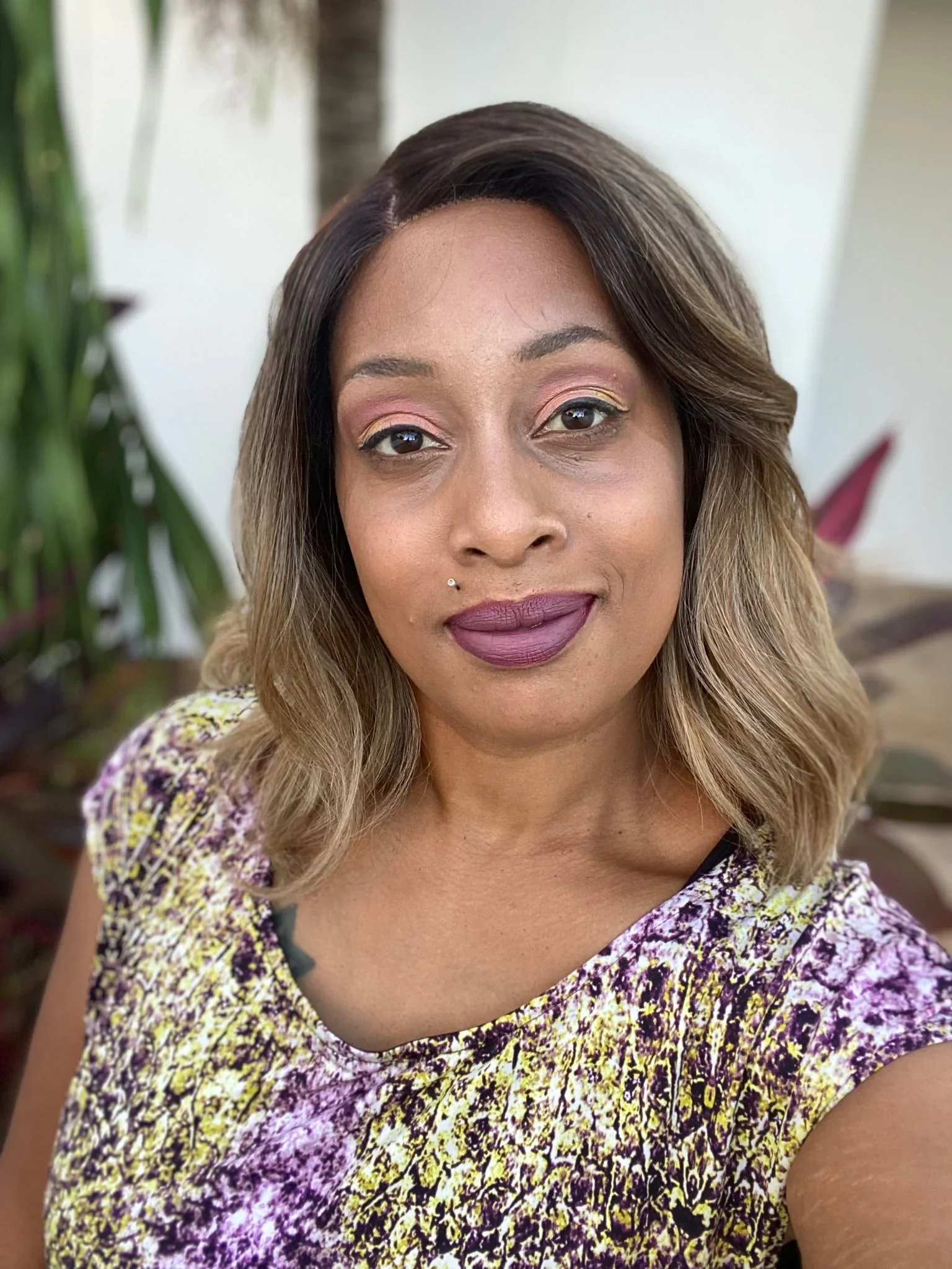 A woman with long wavy hair, wearing makeup with pink and gold eyeshadow and mauve lipstick, smiling slightly, wearing a purple, yellow, and black patterned top, with green plants in the background.