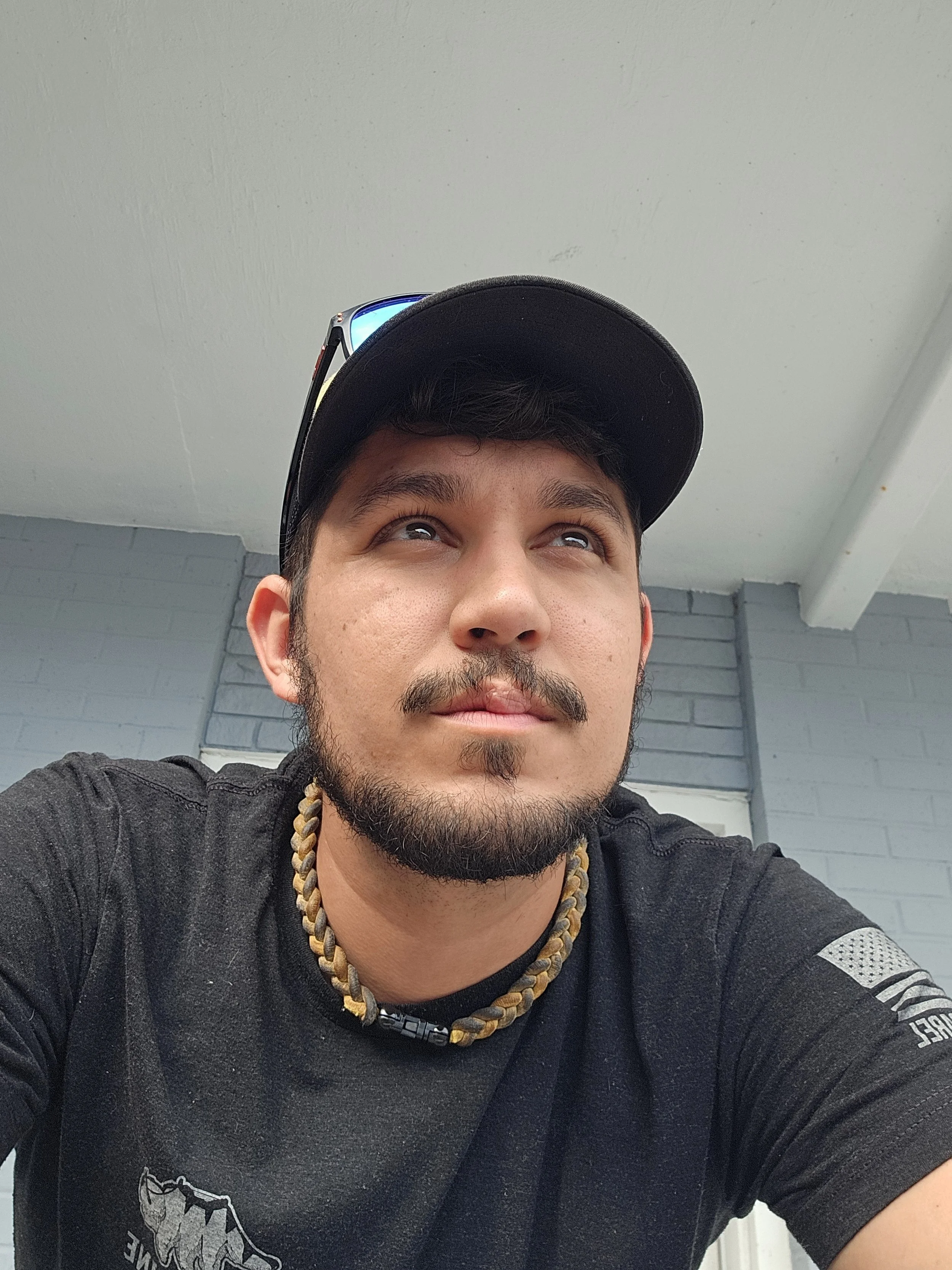 A young man with a beard and mustache wearing a black shirt, a black cap with sunglasses on top, a beaded necklace, and a patch on his left sleeve. He is looking upward with a neutral expression, and the background shows a gray brick wall and a white ceiling.