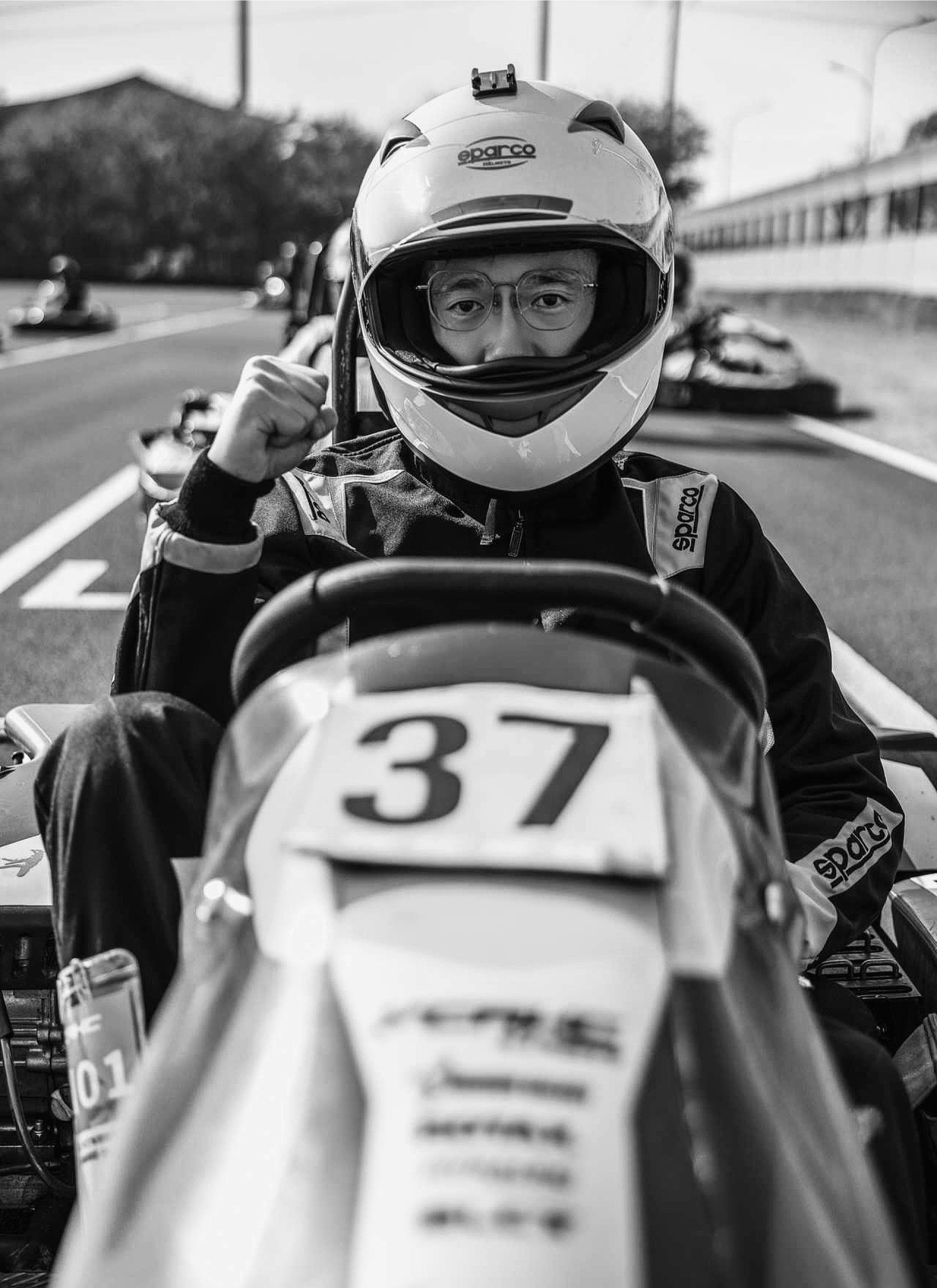 A race car driver in a helmet and racing suit sitting in a go-kart with the number 37, raising a fist in victory or confidence, on a race track with other go-karts in the background.