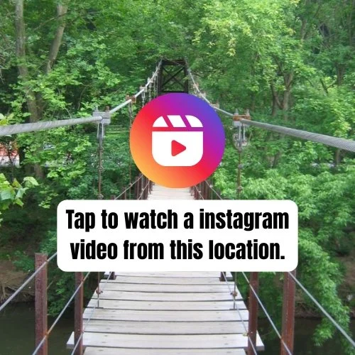 Suspension bridge surrounded by green trees over a forest stream, with a colorful Instagram logo and text overlay prompting to watch a video from this location.
