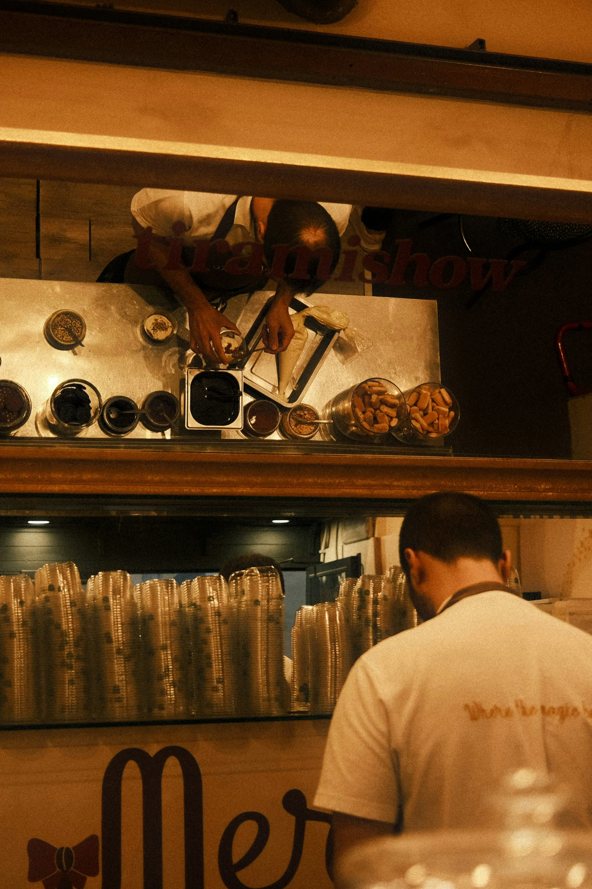 An overhead view of a barista making drinks behind a counter, with plastic cups stacked in front. The barista is working with various ingredients and utensils, seen from below through a window, with a customer standing at the counter.