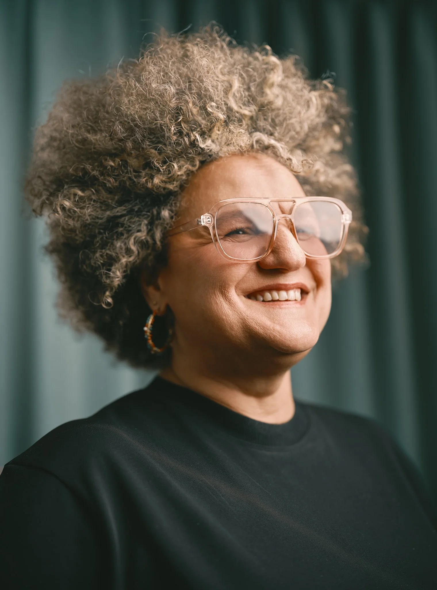 A smiling woman with curly, gray hair wearing large clear glasses, hoop earrings, and a black top, standing in front of a green curtain.