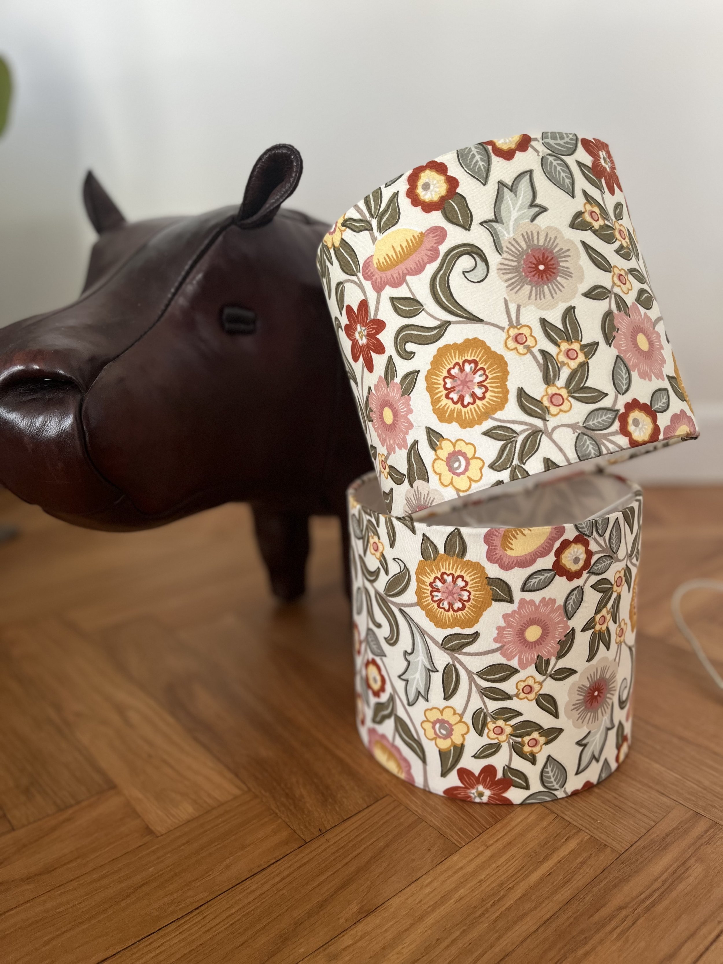 A leather hippo-shaped footstool next to a floral patterned lampshade on a wooden floor.