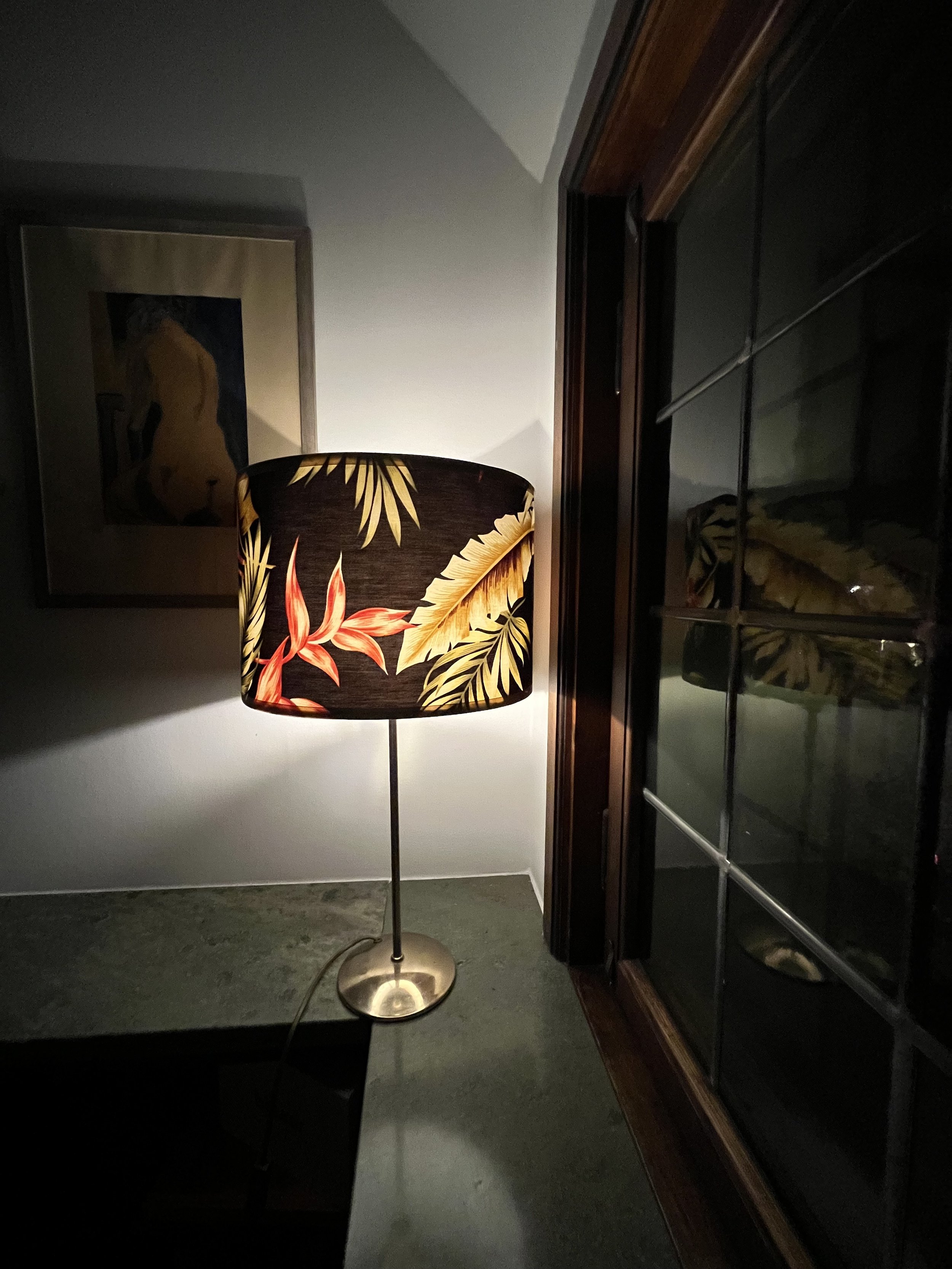 A lamp with a tropical-themed lampshade on a table next to a window, casting shadows on the wall and illuminating a framed artwork.