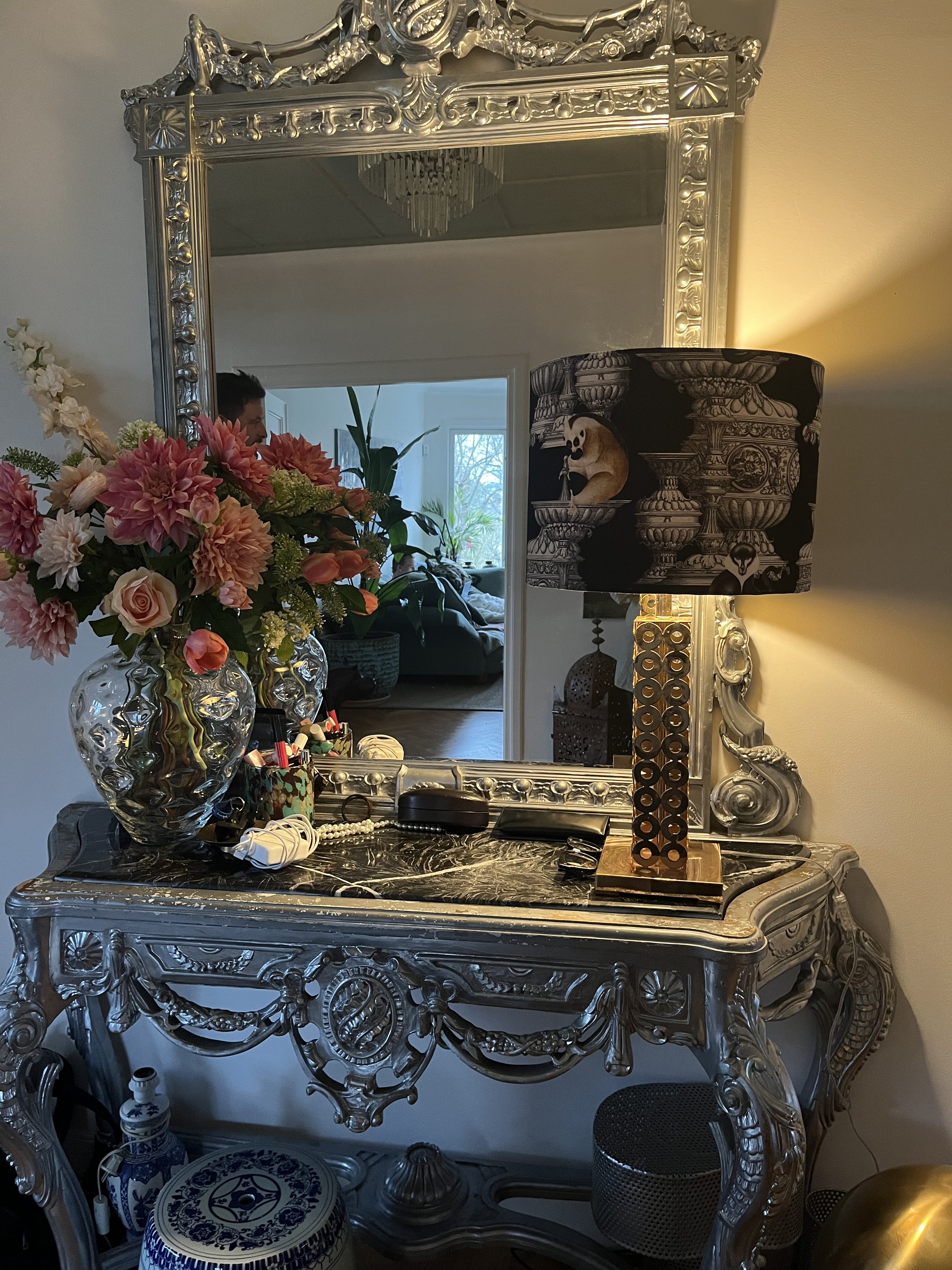 Ornate silver console table with a large decorative mirror and floral arrangement, featuring a patterned lampshade and personal items on the table surface in a living room setting.