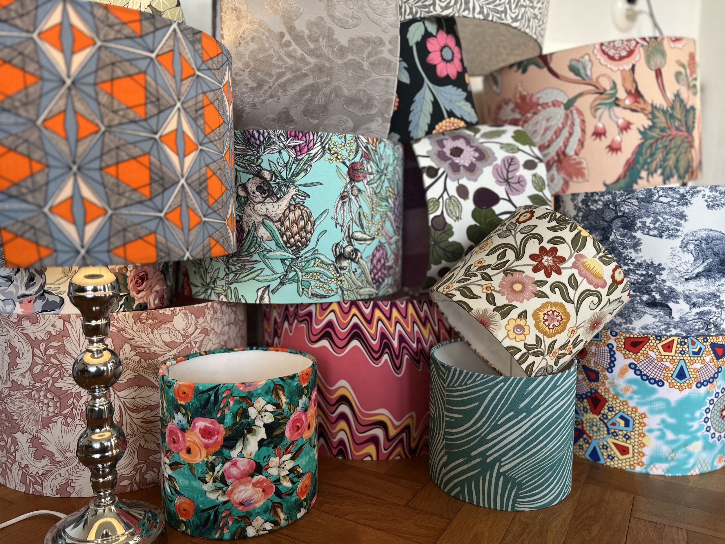 A collection of colorful and patterned lampshades, including floral, geometric, and abstract designs, with a silver lamp base partially visible.