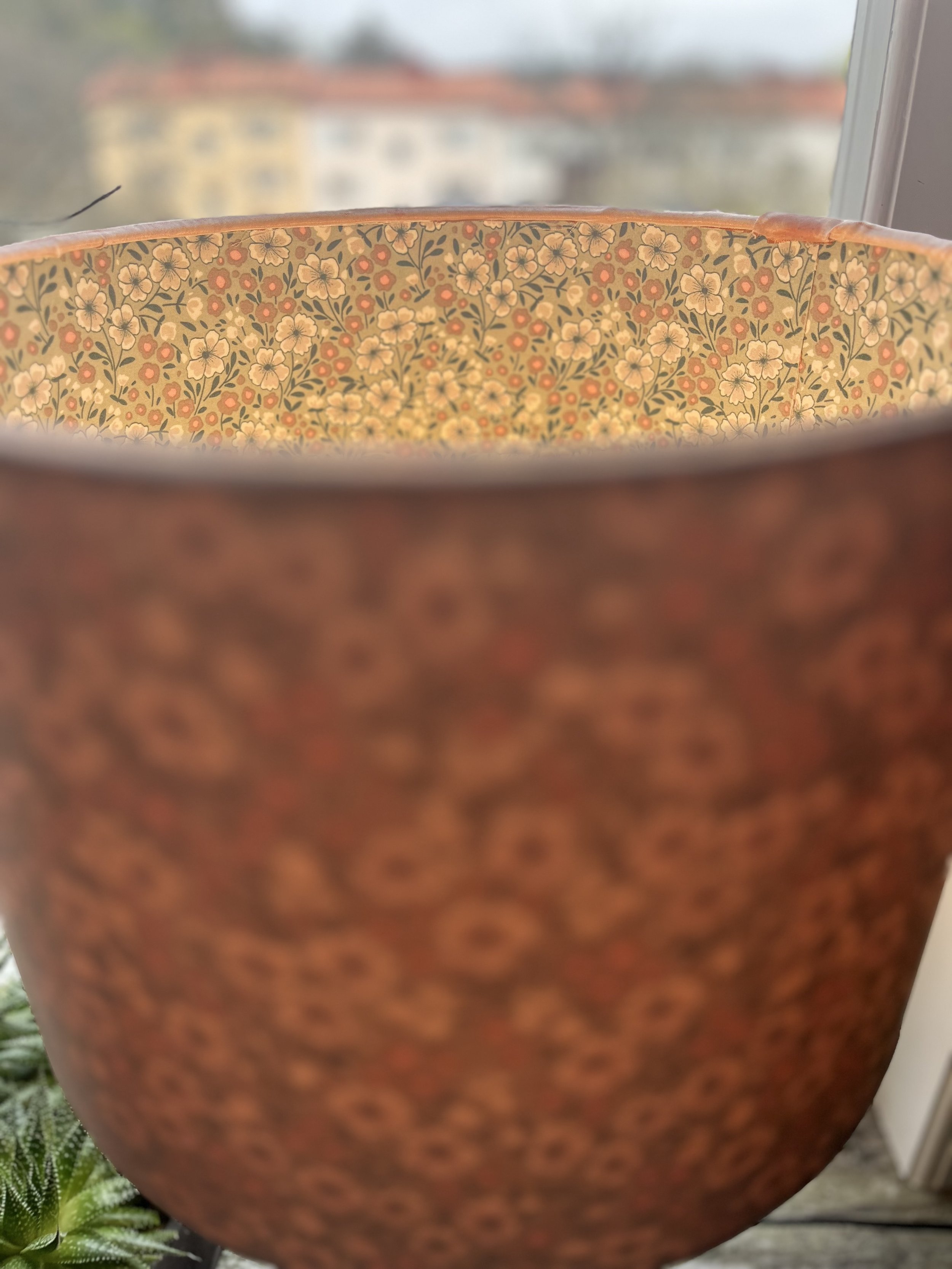 Close-up of a ceramic flower pot with a floral pattern, viewed from the top, with blurred background of buildings and greenery outside a window.