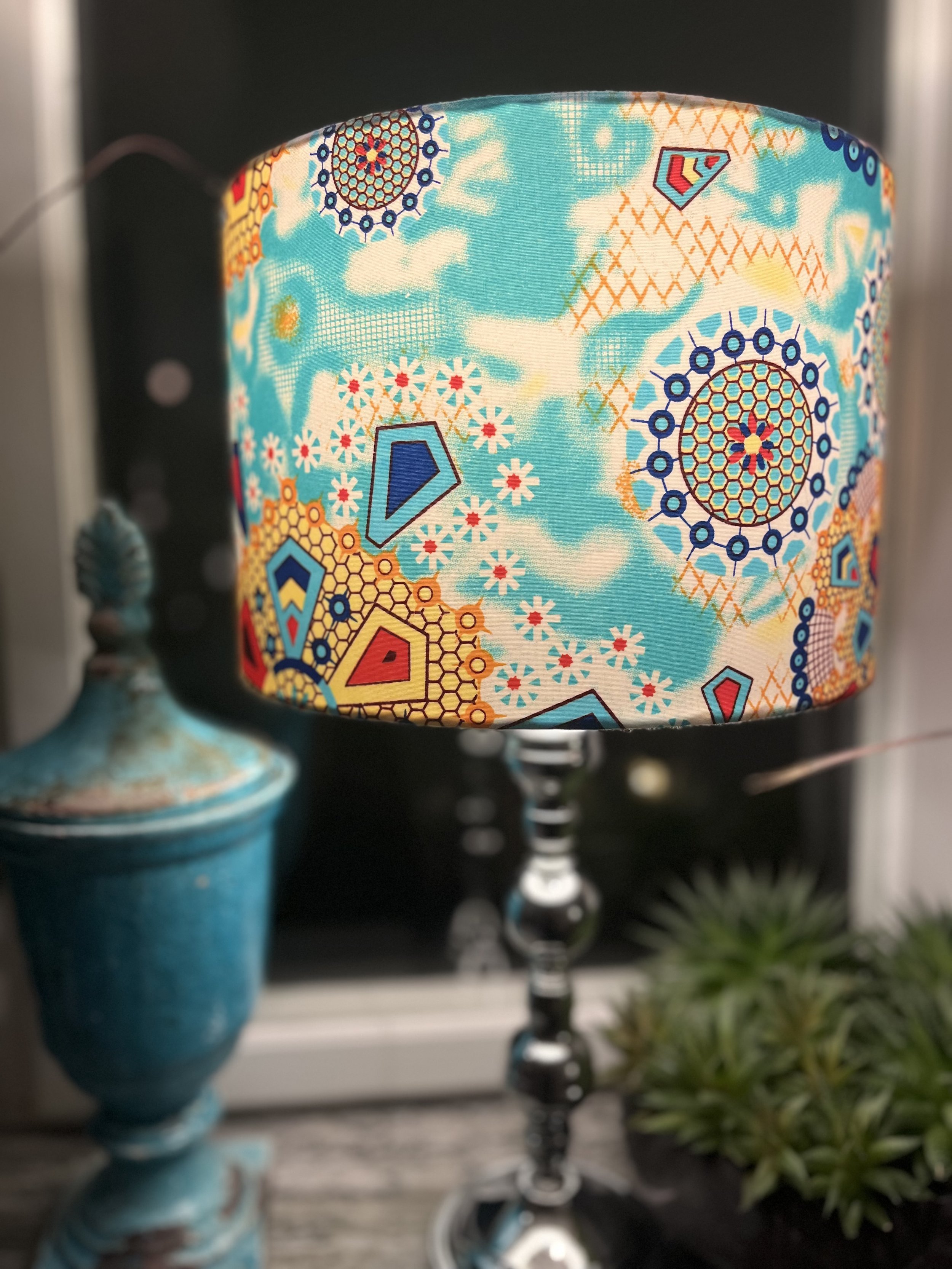 Decorative lamp with colorful patterned lampshade and silver base next to a blue vase and potted succulents on a windowsill.