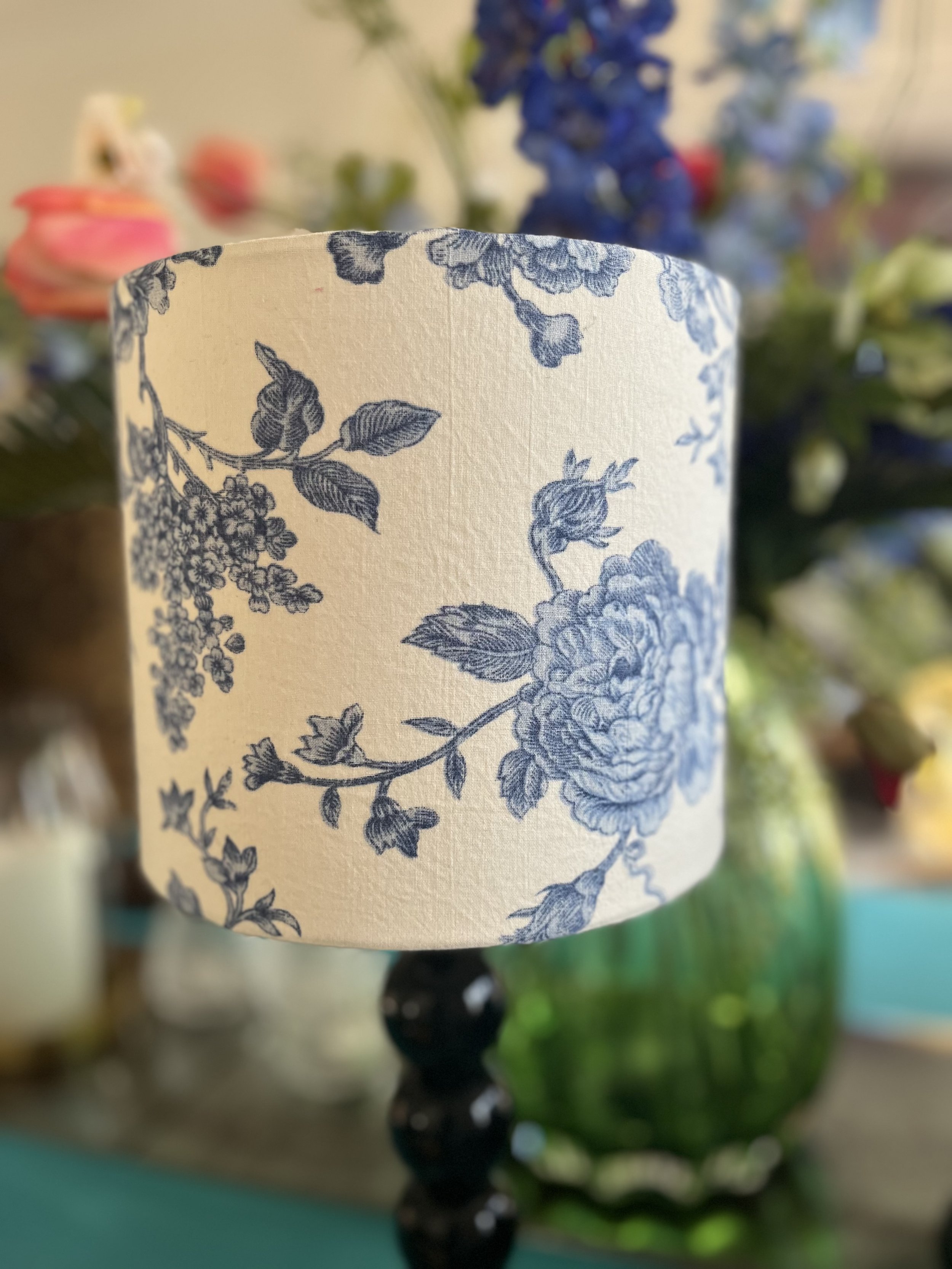 Close-up of a lampshade with blue floral pattern, set against a blurry background of colorful flowers and a teal surface.