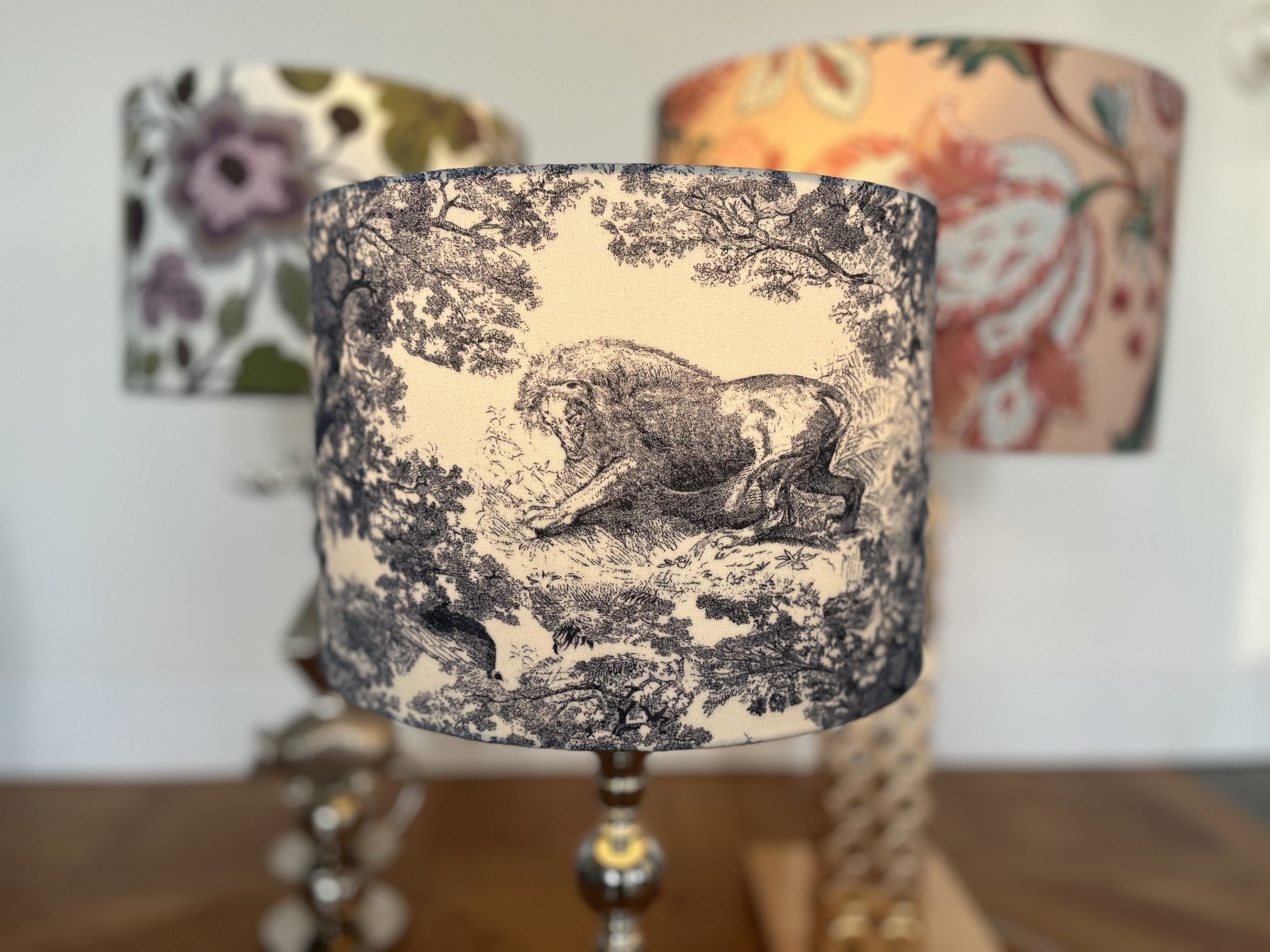 Three decorative lampshades with intricate designs; the foreground features a toile pattern with a lion and trees, while the background shows floral patterns in various colors.