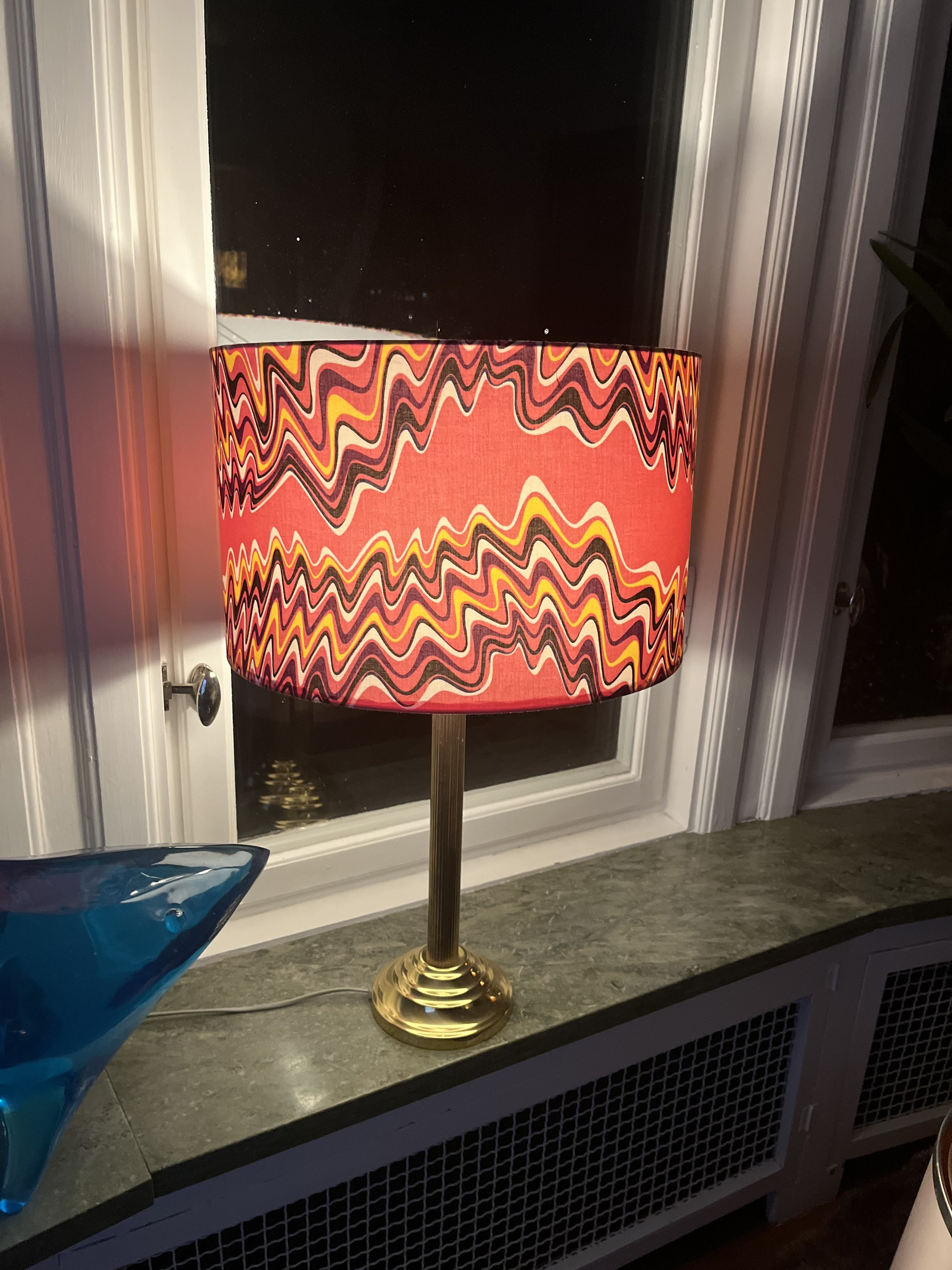 Table lamp with a vibrant, wavy-patterned lampshade in red, yellow, and black colors on a window sill next to a blue glass decor object.