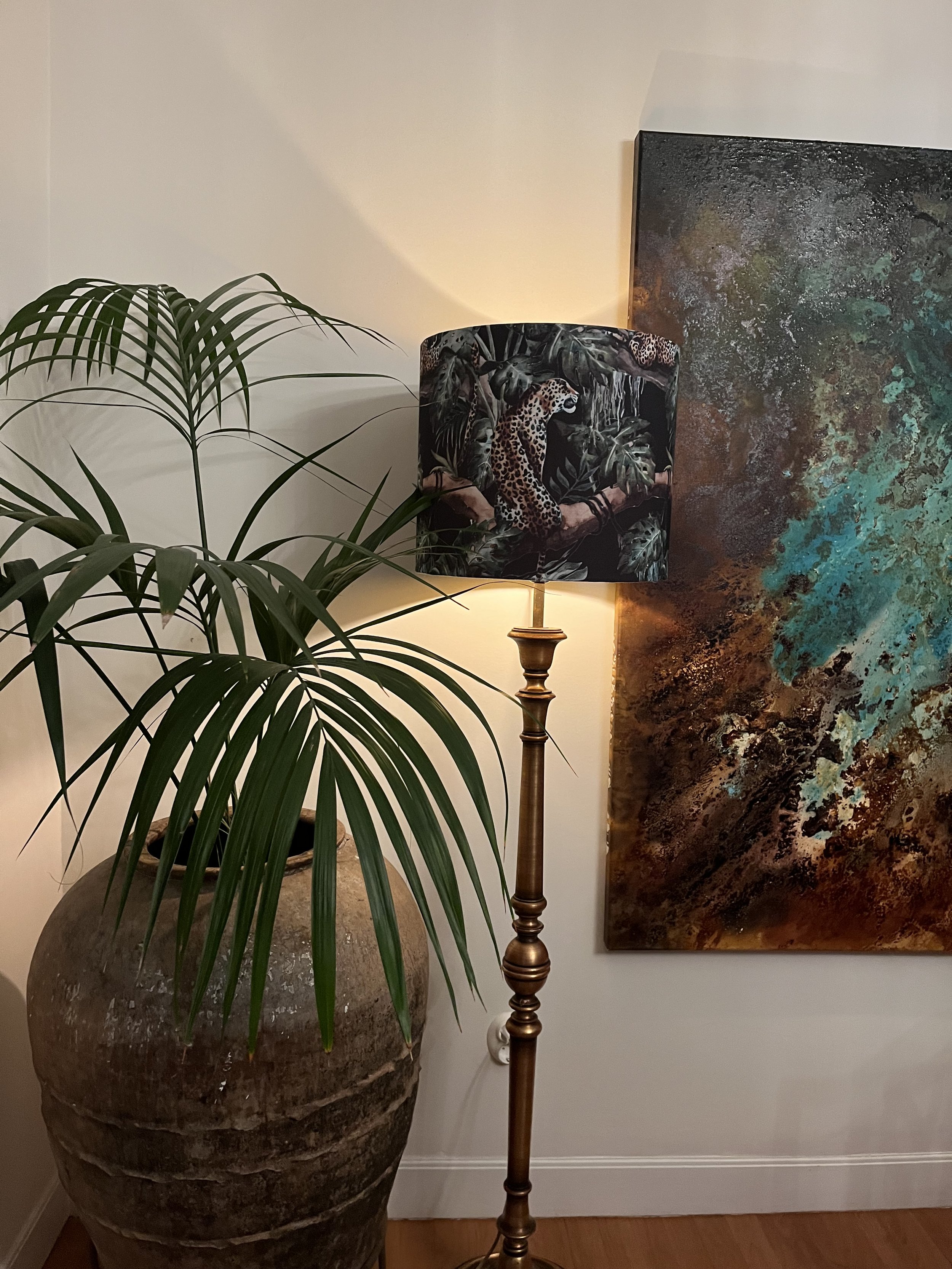 A room corner with a large potted plant in a textured vase next to a floor lamp. The lamp has a shade featuring a leopard design. An abstract painting with blue and brown tones hangs on the wall nearby.