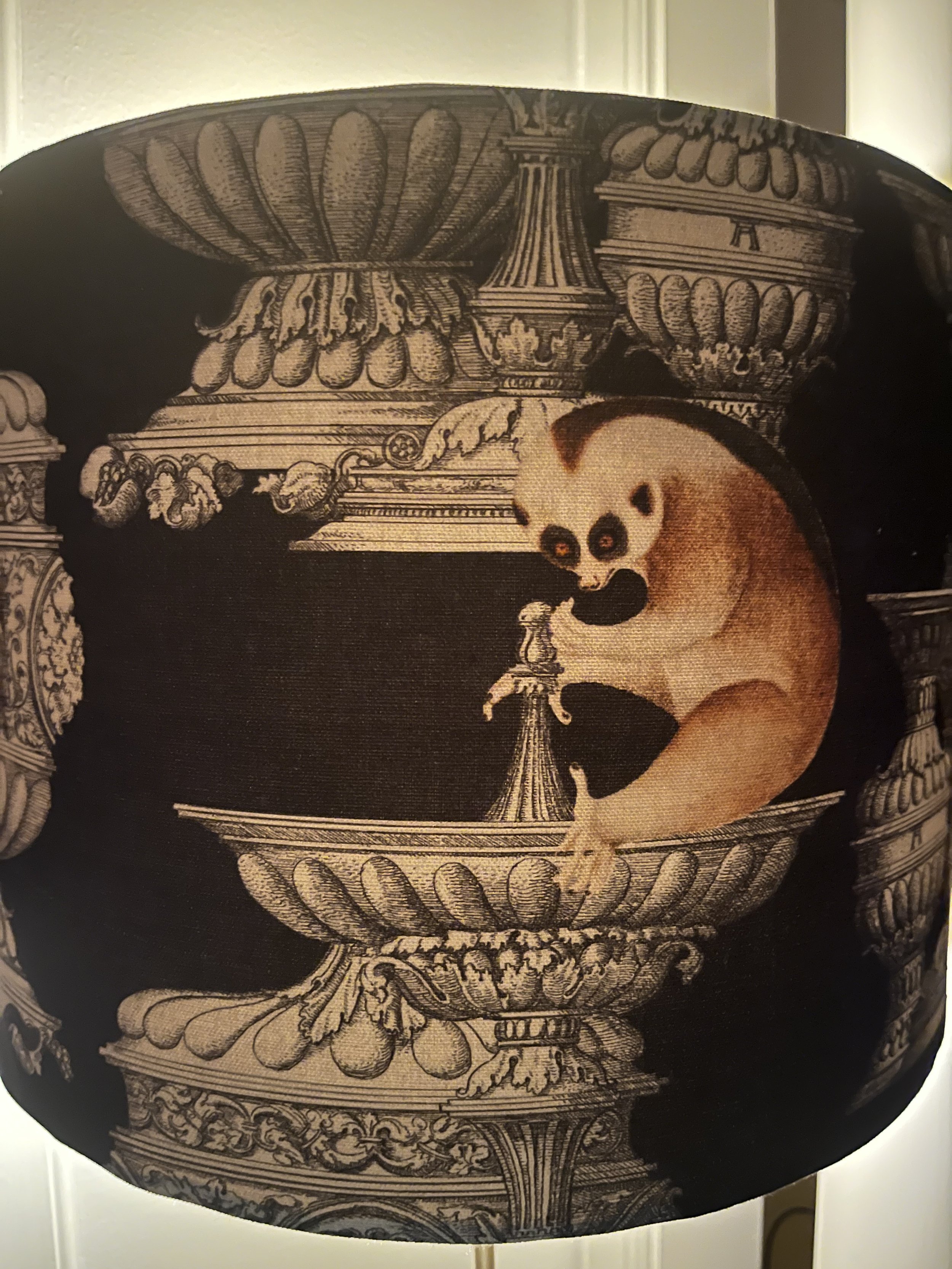 Decorative lamp shade with intricate architectural designs and a small animal illustration, featuring detailed urns and pillars on a dark background.