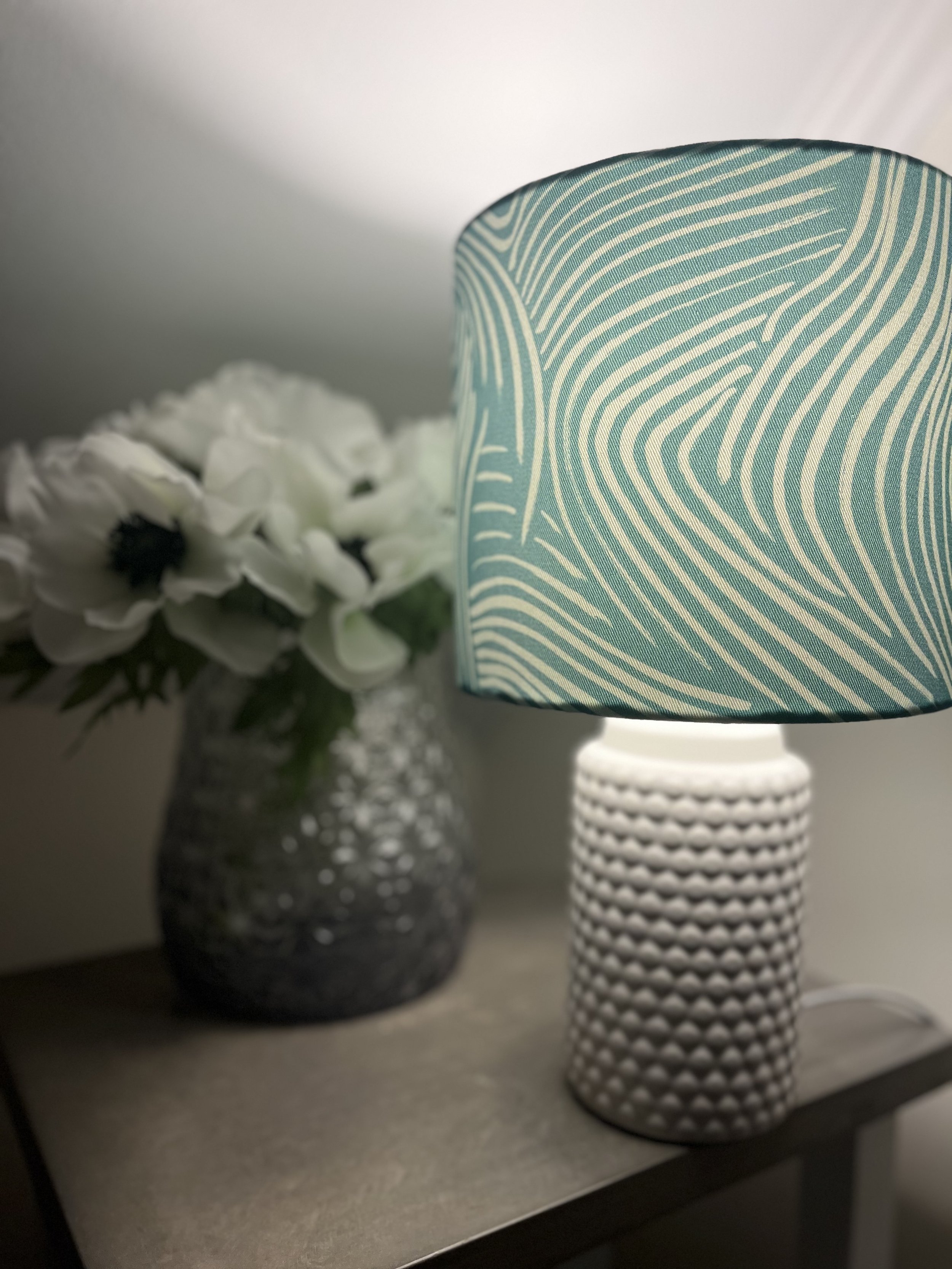 Table with a decorative lamp featuring a teal-patterned shade and a textured white base, next to a vase of white flowers.