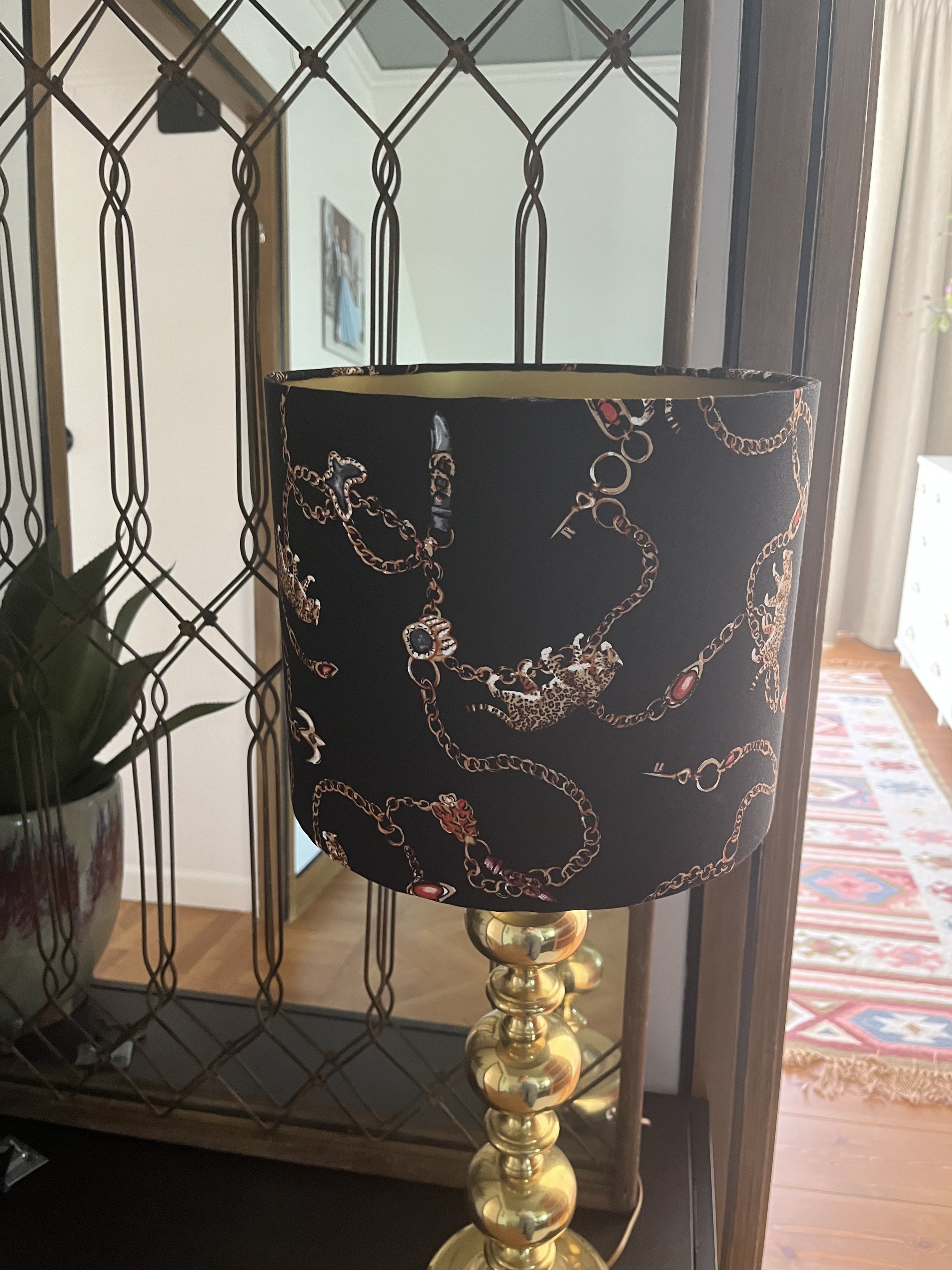 Decorative lamp with a patterned black lampshade featuring gold chain designs, set on a brass base, placed in front of a mirror with a decorative metal frame, next to a plant pot on a dark surface.