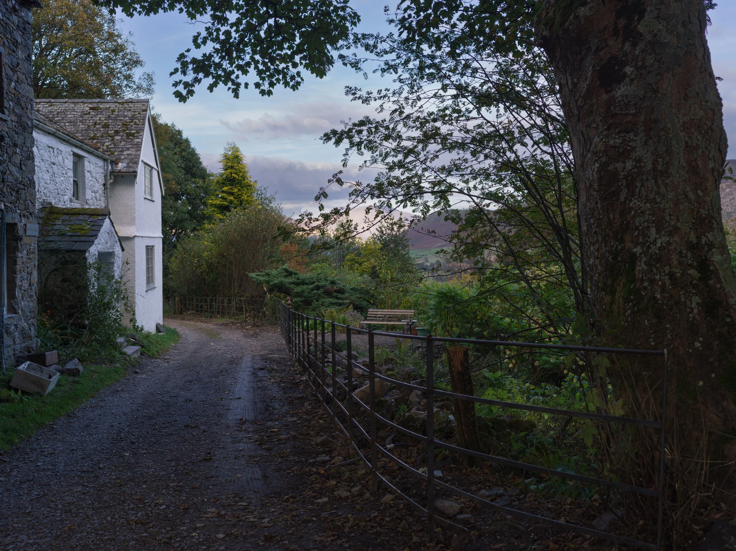 A dirt path runs alongside a stone house with white walls and a tiled roof. A metal railing lines the path on the right, with trees and greenery beyond. A bench is visible in the background, and the sky is partly cloudy during what appears to be late