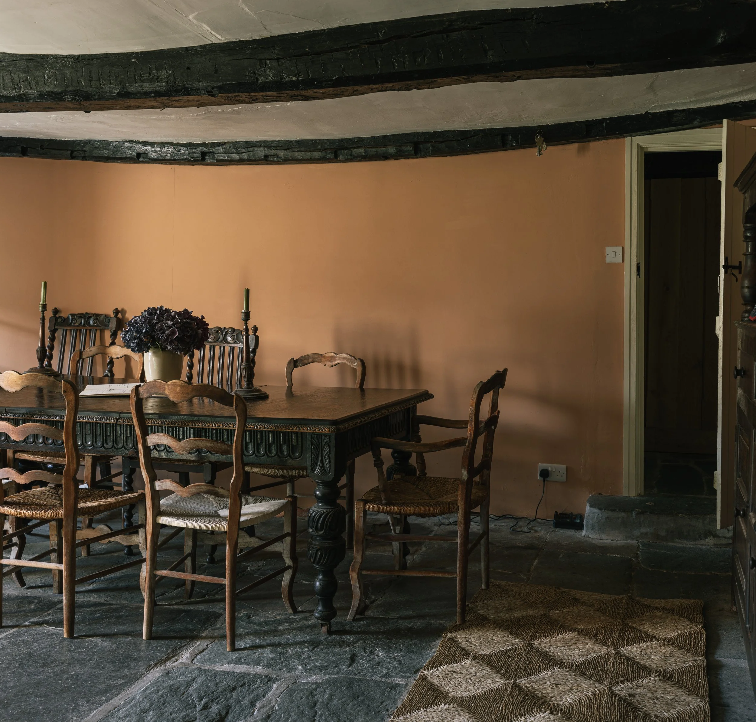 Interior of a dining room with a wooden table, six chairs, a beige wall, a vase with dark purple flowers, and two green candlesticks. The floor is stone, and a doorway leads to a dark room.