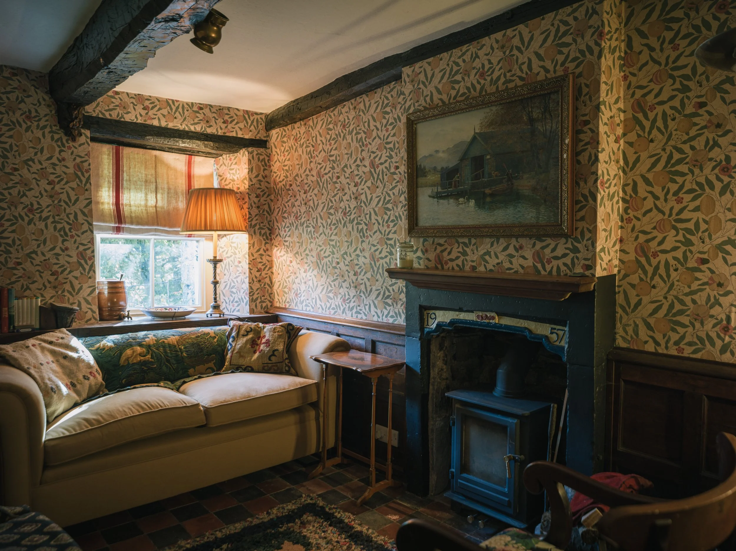 A cozy living room with patterned wallpaper, a beige sofa with floral cushions, a window with a striped curtain, a table lamp, a fireplace, a framed landscape painting, and rustic wooden beams.