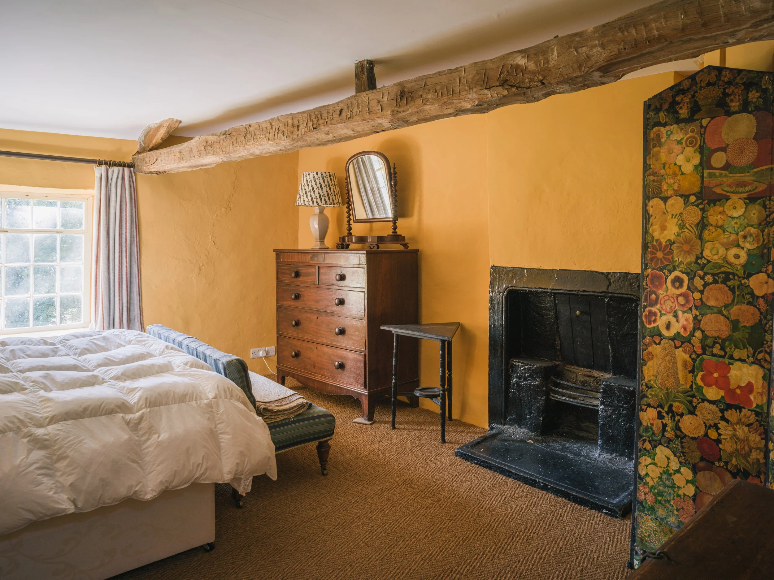 A cozy bedroom with yellow walls, a window with striped curtains, a bed with white bedding, a wooden dresser with a mirror and lamp, a small black side table, a black fireplace, and a floral room divider.