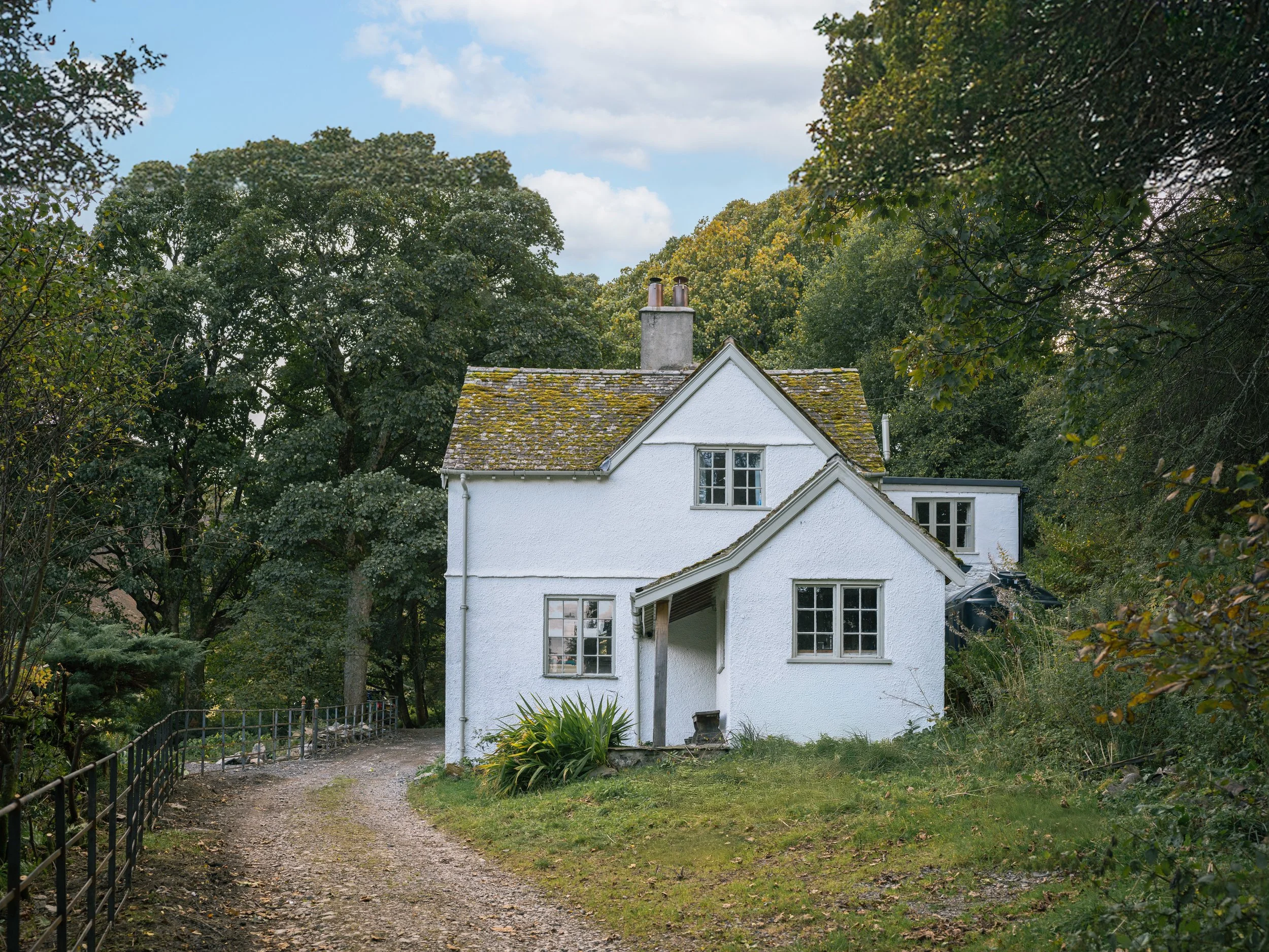 A white house with a sloped roof surrounded by trees and greenery, with a gravel pathway leading to the front door.
