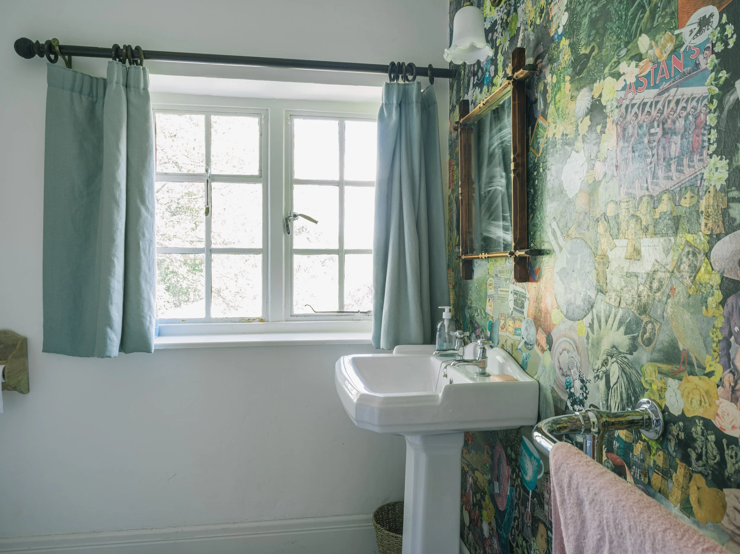 A bathroom with light blue curtains, a window, a vintage mirror, a white sink with soap and sanitizer bottles, a towel hanging on a chrome bar, and a colorful collage mural of various images on the wall.