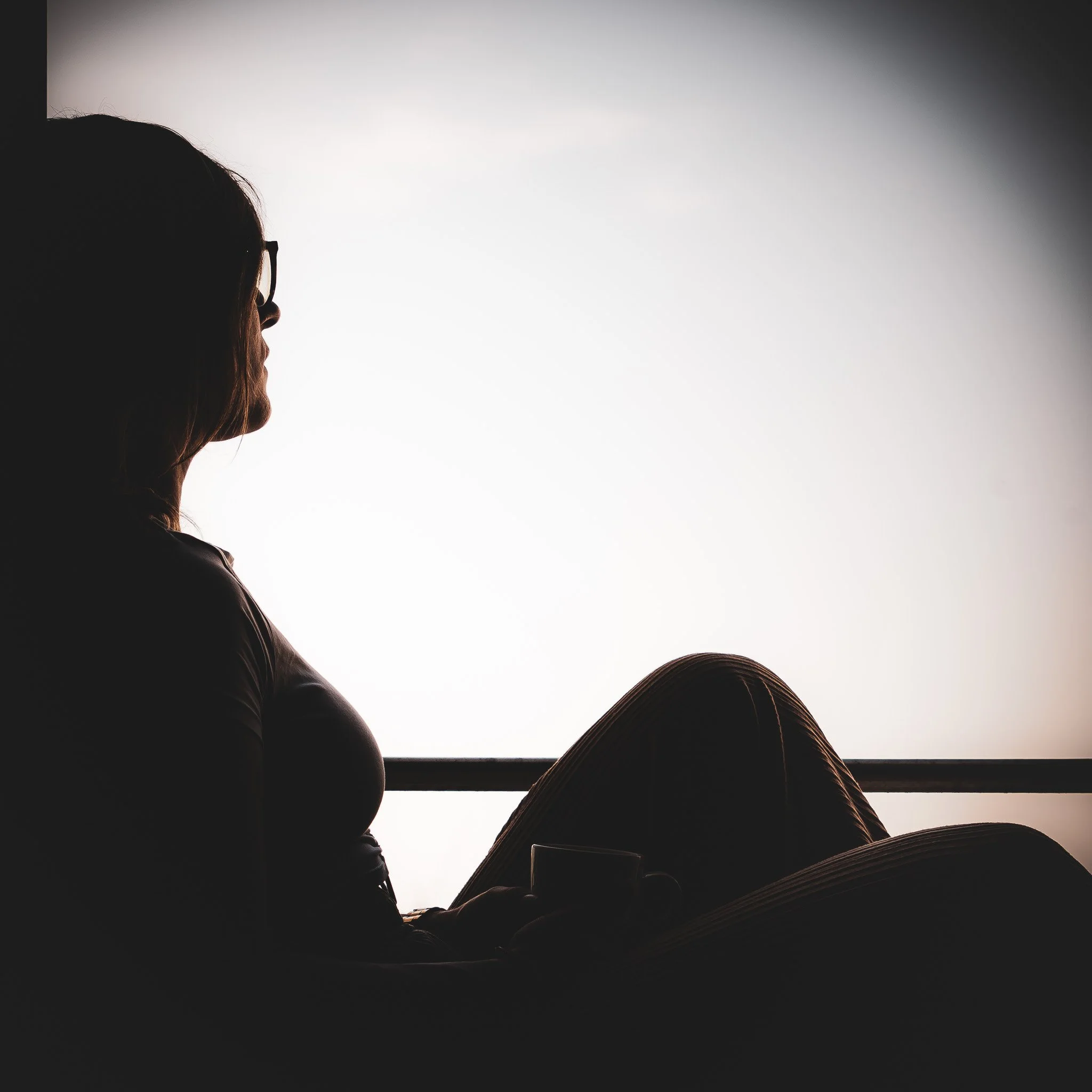 Silhouette einer Person mit Brille, die an ein Fenster gelehnt sitzt, mit einer Tasse in der Hand.