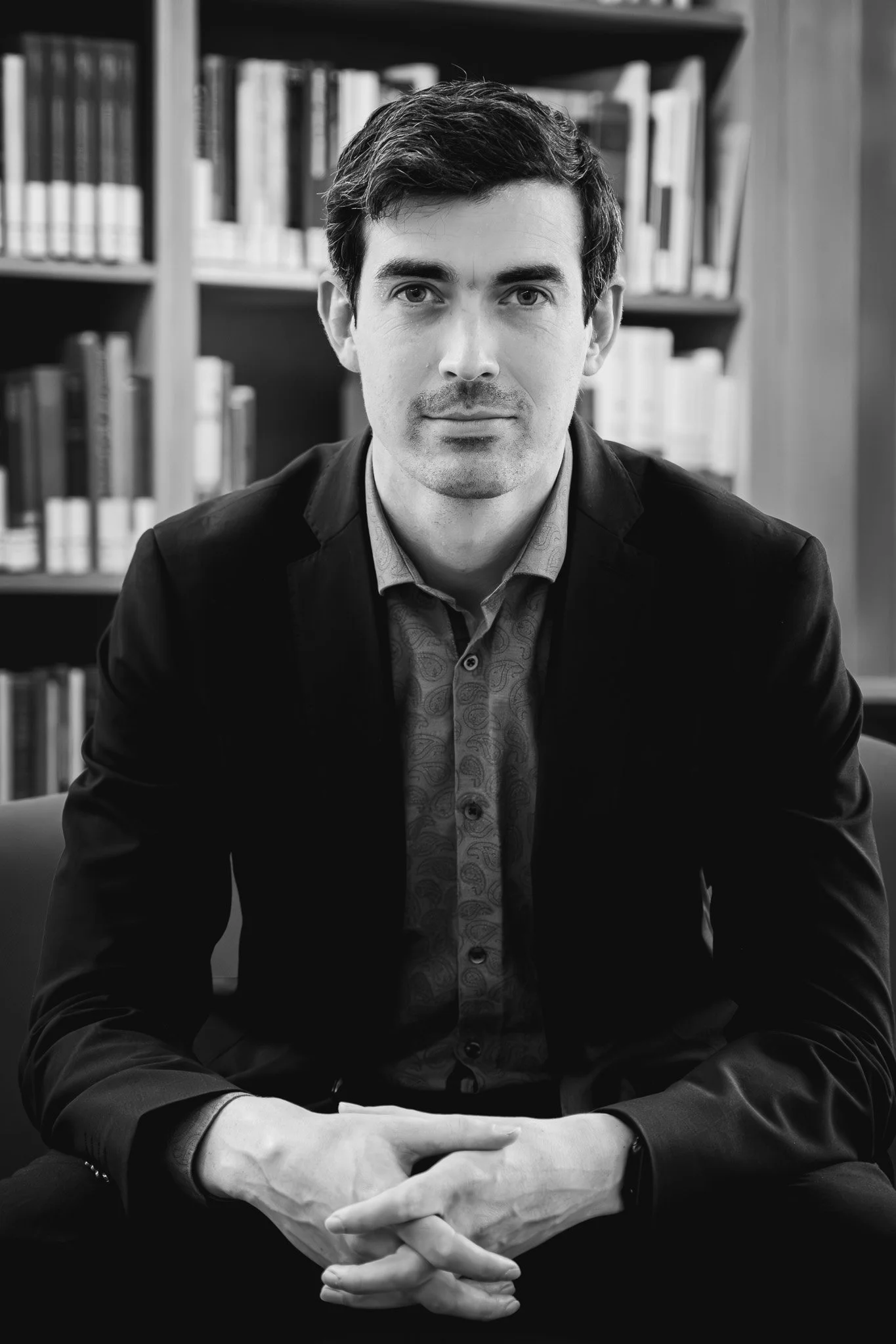 Schwarz-weiß-Foto eines Mannes im Anzug, der vor einem Bücherregal sitzt.