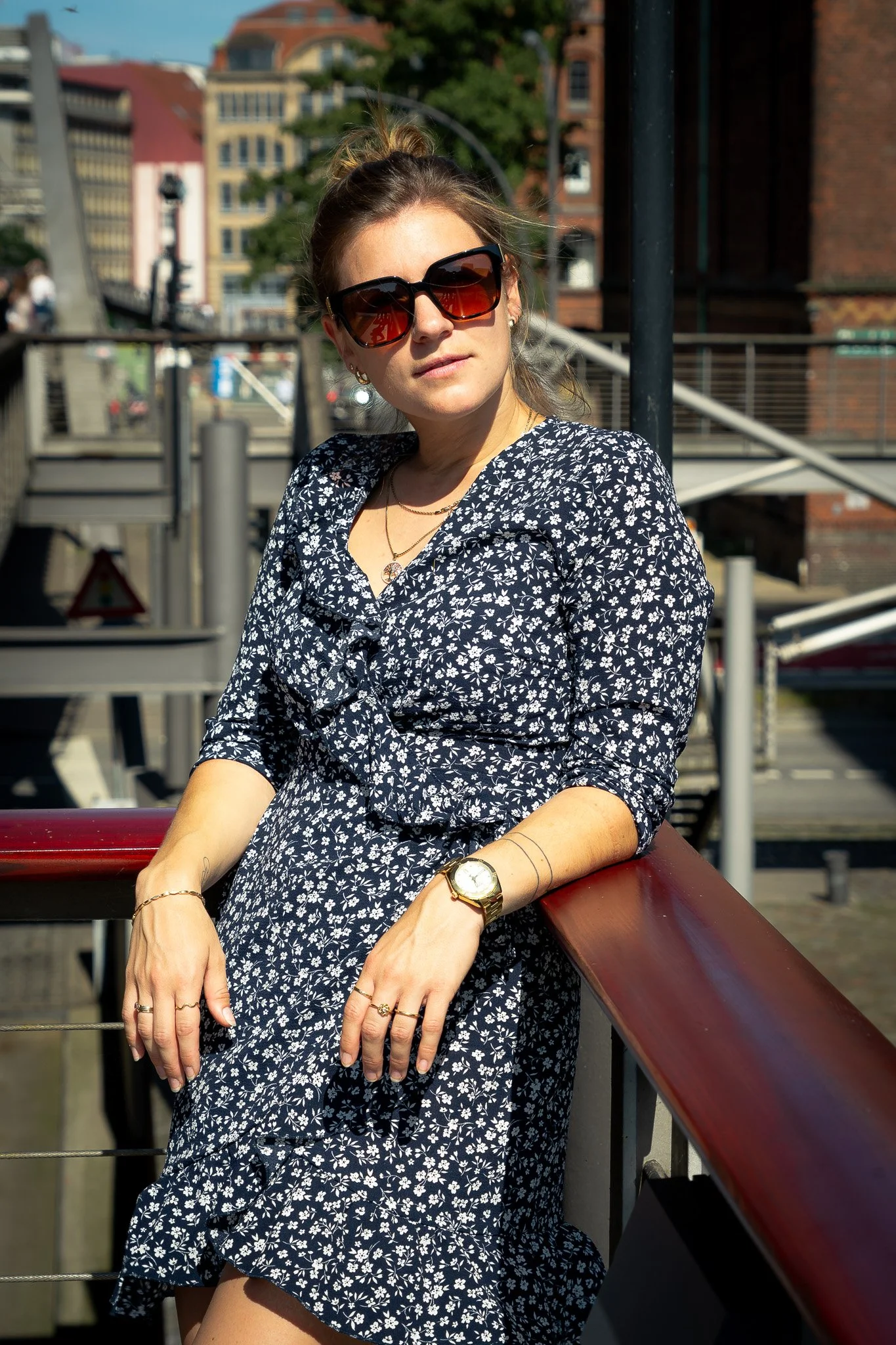 Frau mit Sonnenbrille steht im Freien an einem Geländer. Sie trägt ein blaues Kleid mit weißem Blumenmuster."